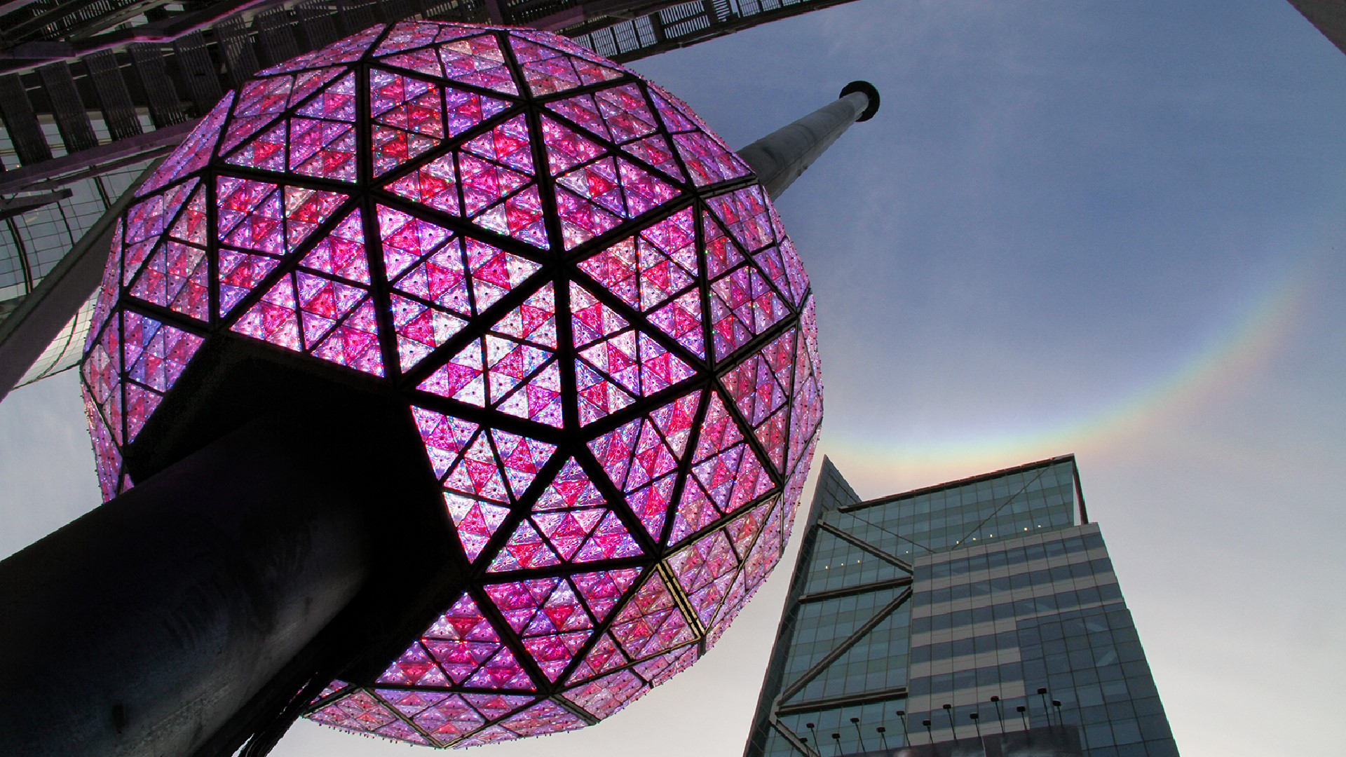 Times Square’s New Year’s Eve ball, glowing pink, waits to be hoisted into the air above the city’s skyscrapers.