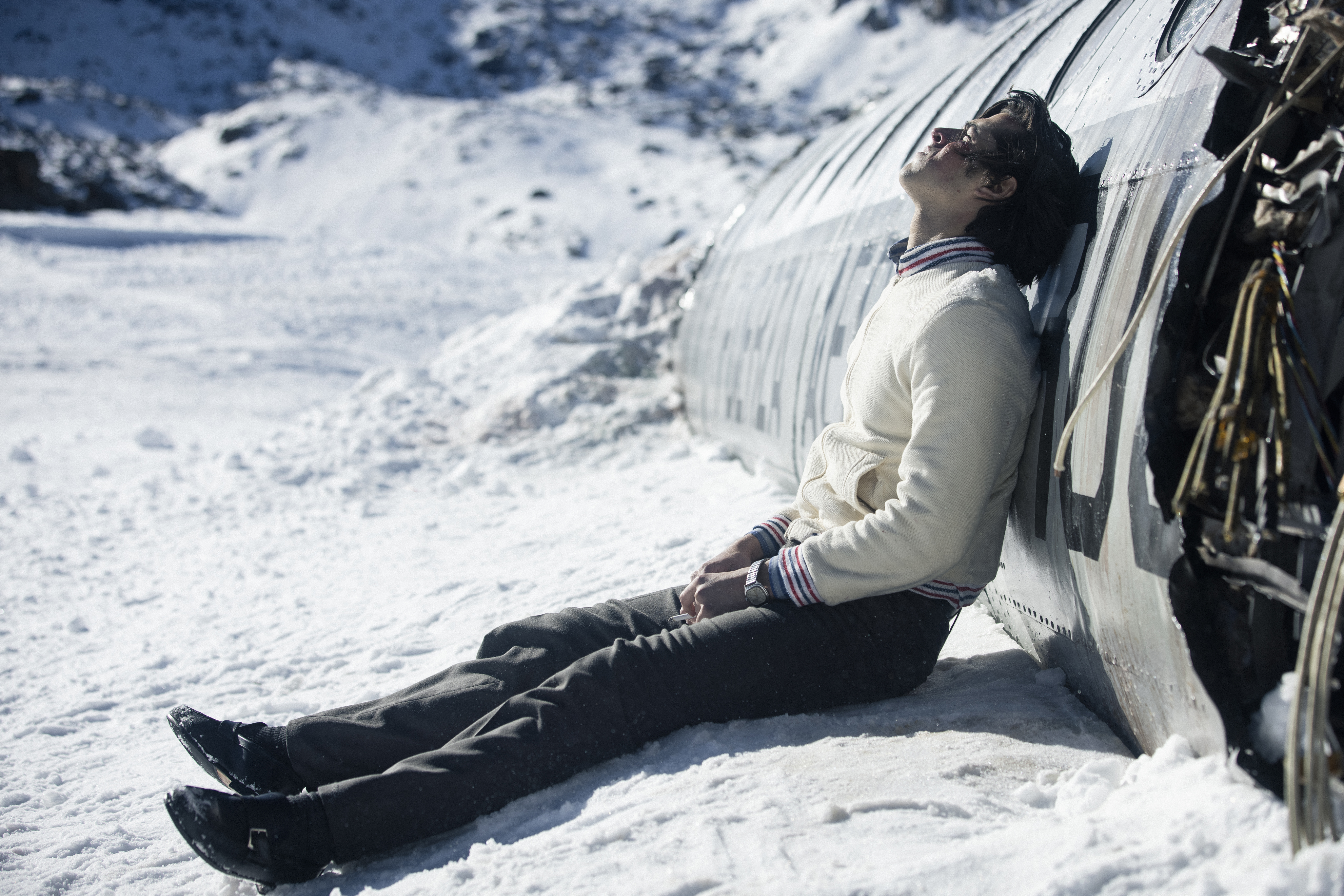 A young man in dark pants and a white sweater, face bruised and bloodied, sits in the snow with his back against the curved side of a torn-open plane in Netflix’s The Society of the Snow
