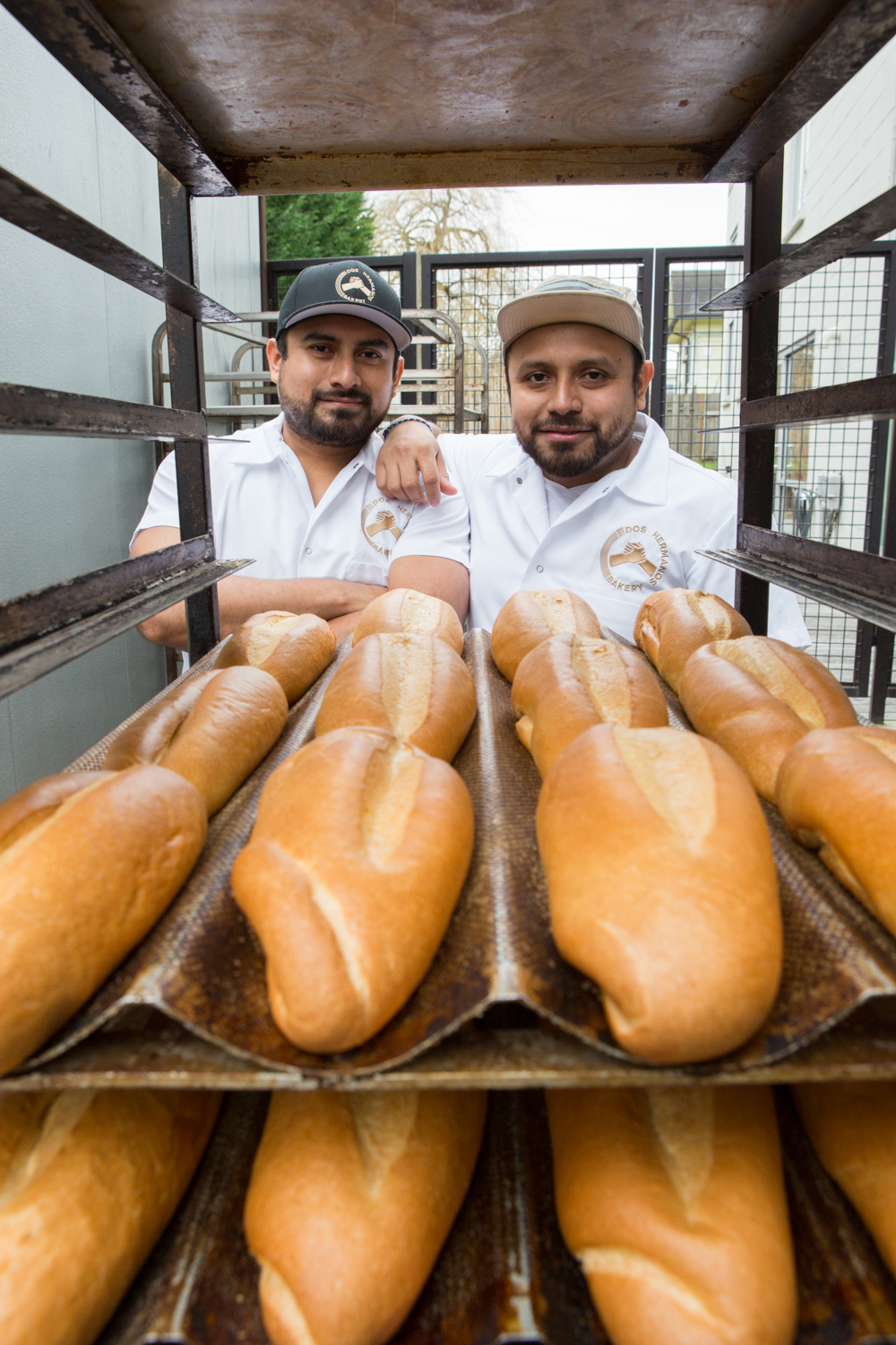 Two men pose on the other side of a rack of fresh bread.