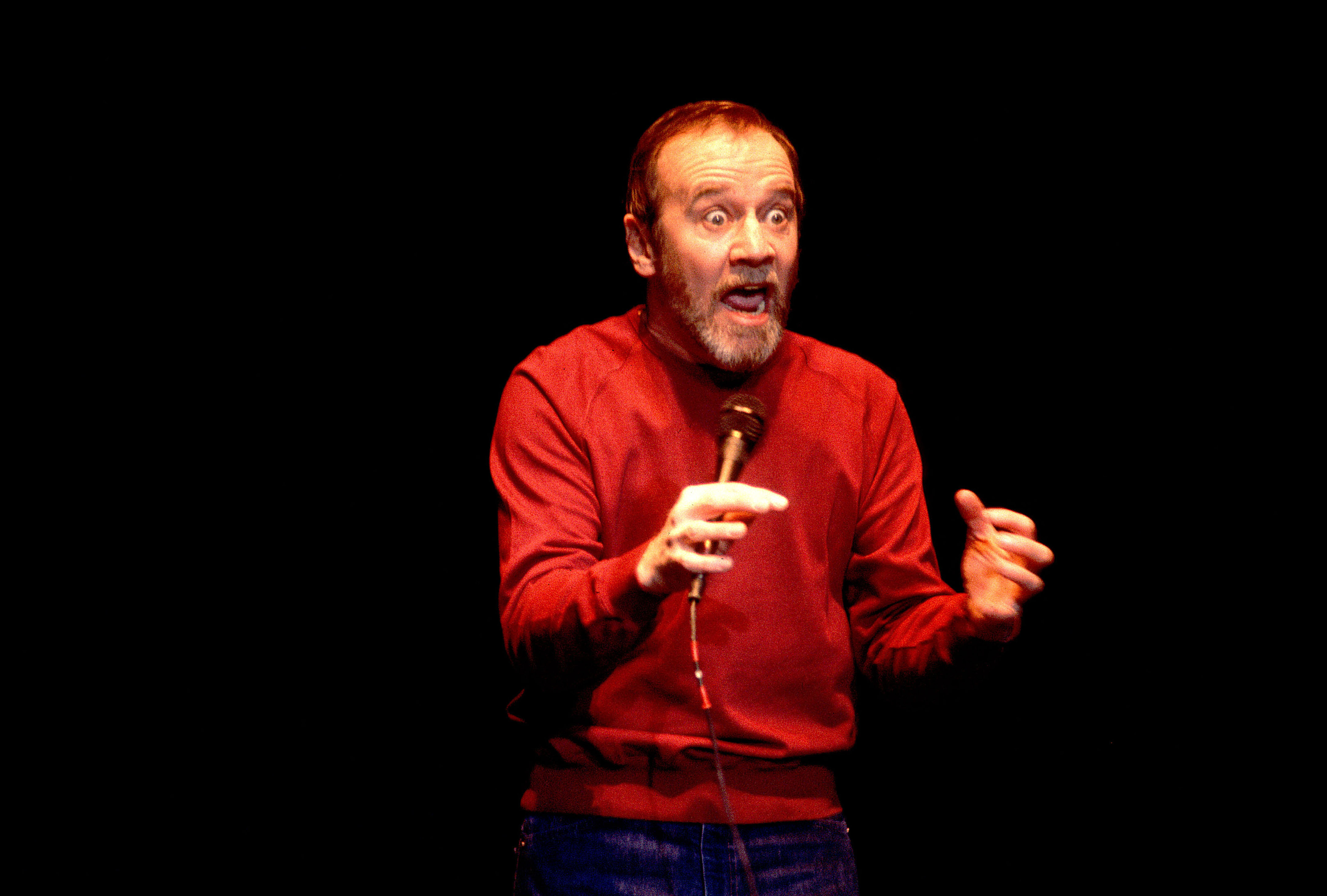 George Carlin performs on-stage. He is brightly lit while the background is black. He is an older white man with grey hair pulled back, a beard, and an exaggerated expression on his face.