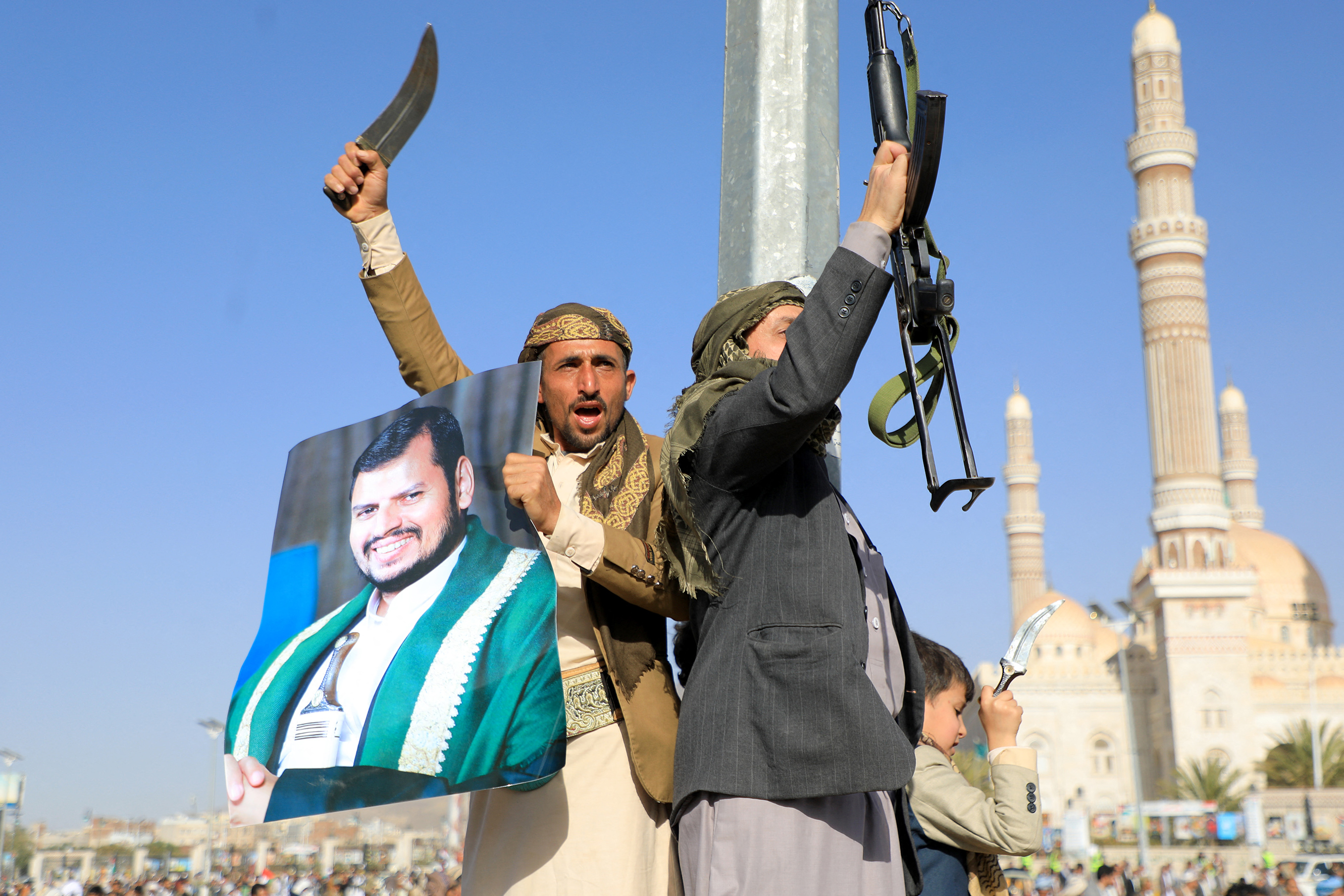 Two men raise weapons against a blue sky: on the left, a man in a tan suit jacket holds a photo of Houthi leader Abdul Malik al-Houthi in one hand and brandishes a curved knife in the other; on the right, a man in a grey suit jacket raises an automatic rifle.