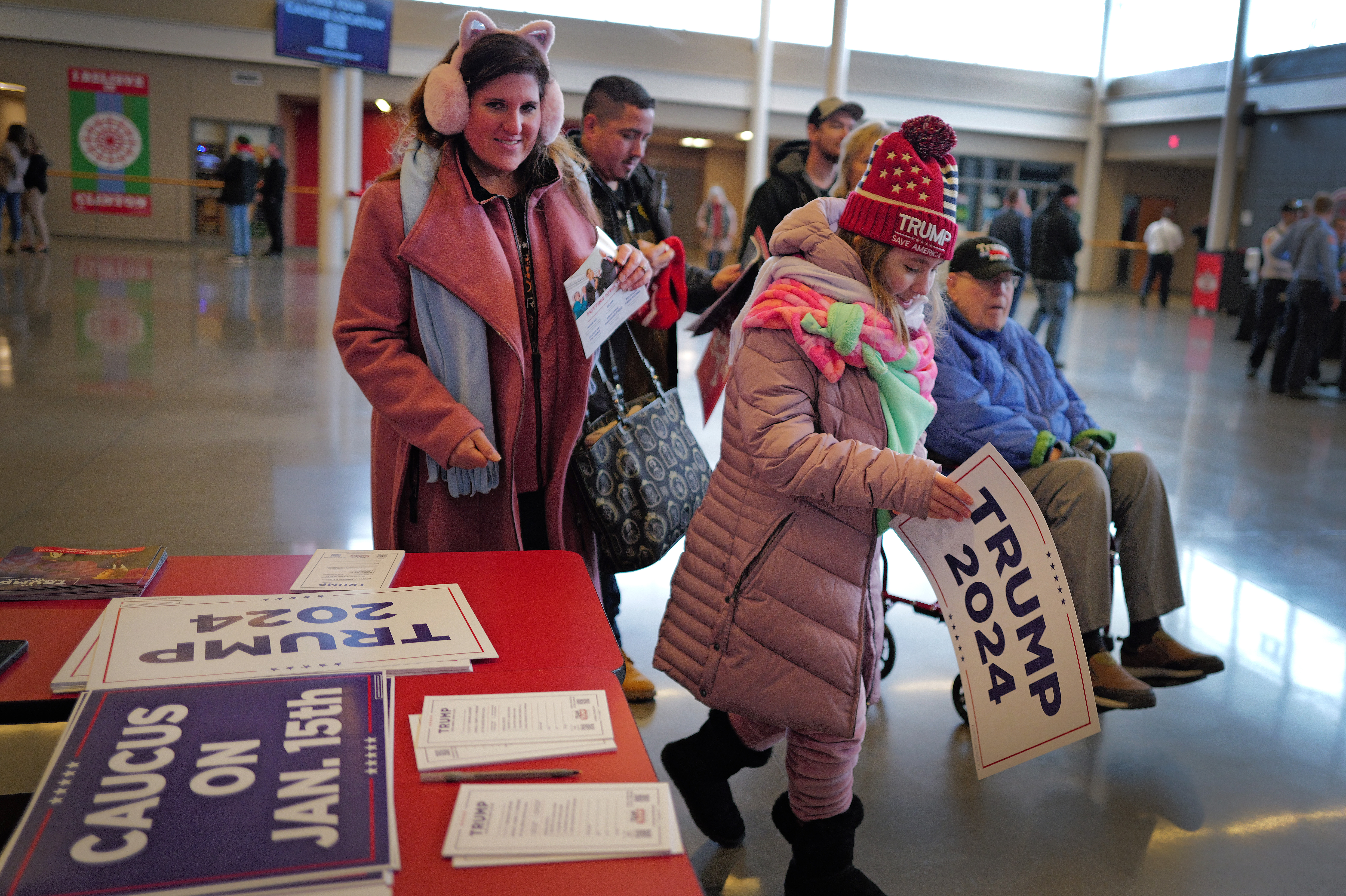 People in winter coats and hats gather in a school building, holding TRUMP 2024 signs and standing next to a table with more signs with political slogans.