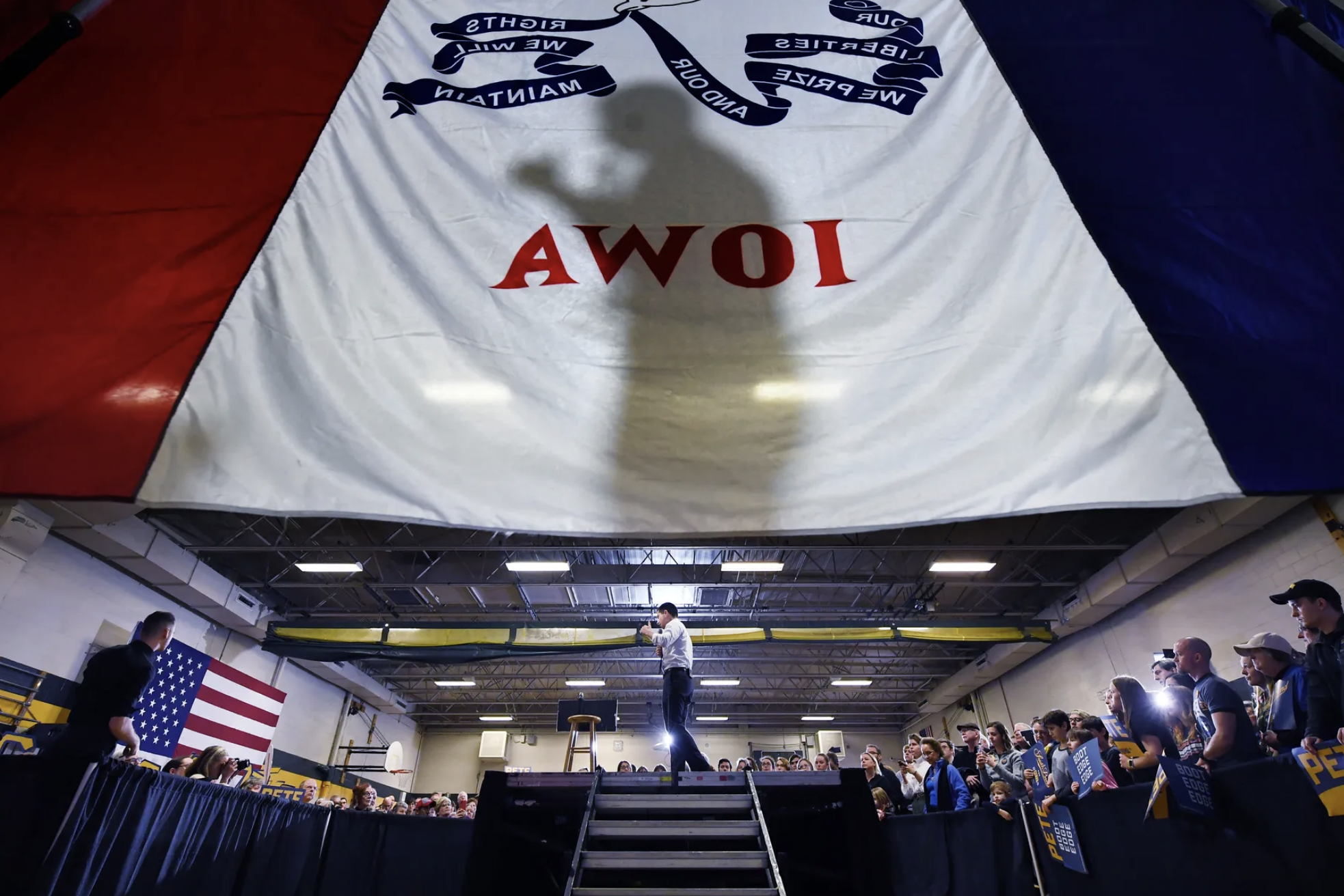 A shadow of a man is cast on a large white banner reading “Iowa” hanging in a school gym, where the middle of the room is a stage platform and the man is walking onto it.
