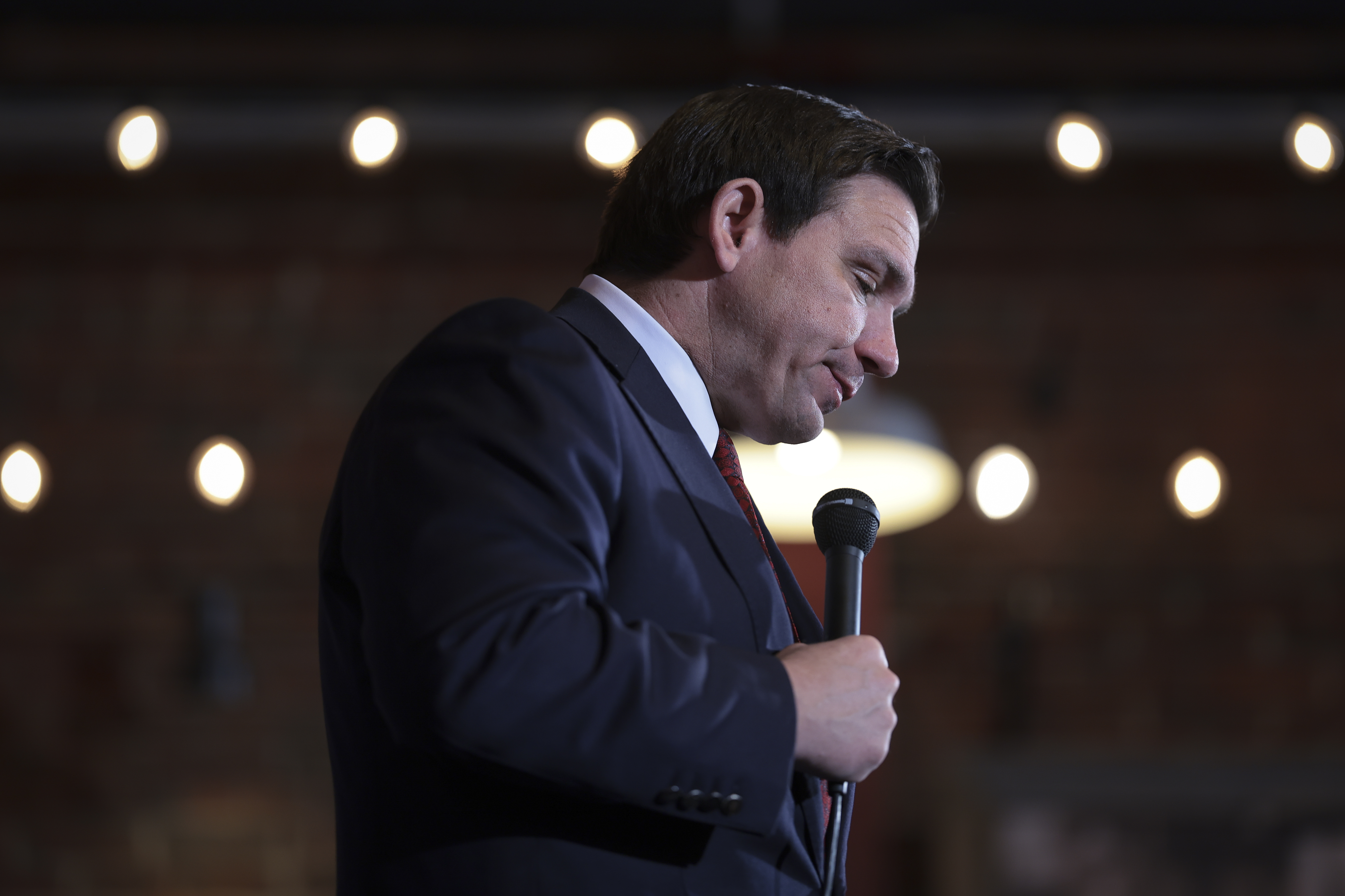 DeSantis in a dark suit against a background of lights, looks down and holds a microphone.