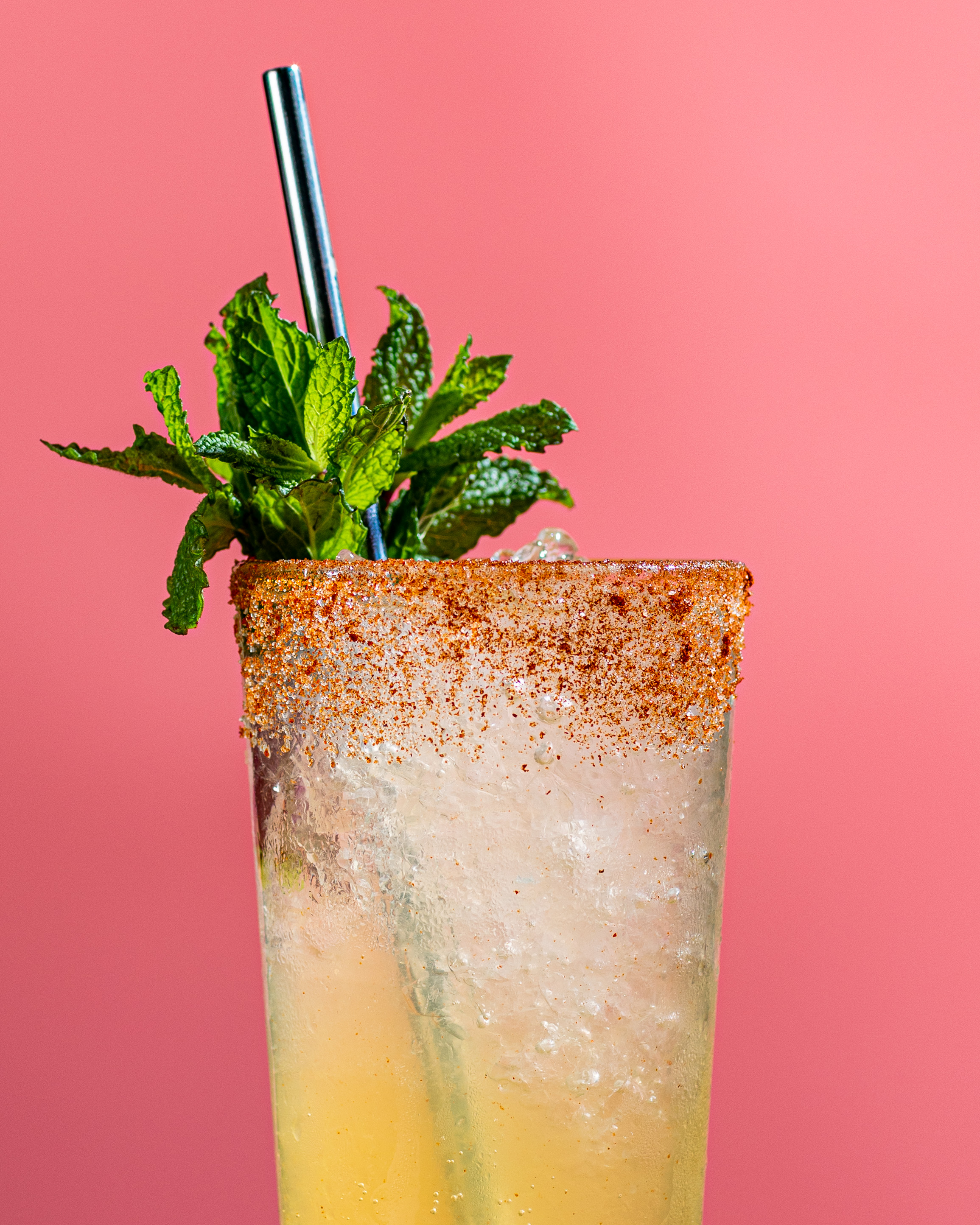 A yellow cocktail drink with a sprig of mint against a pink background.