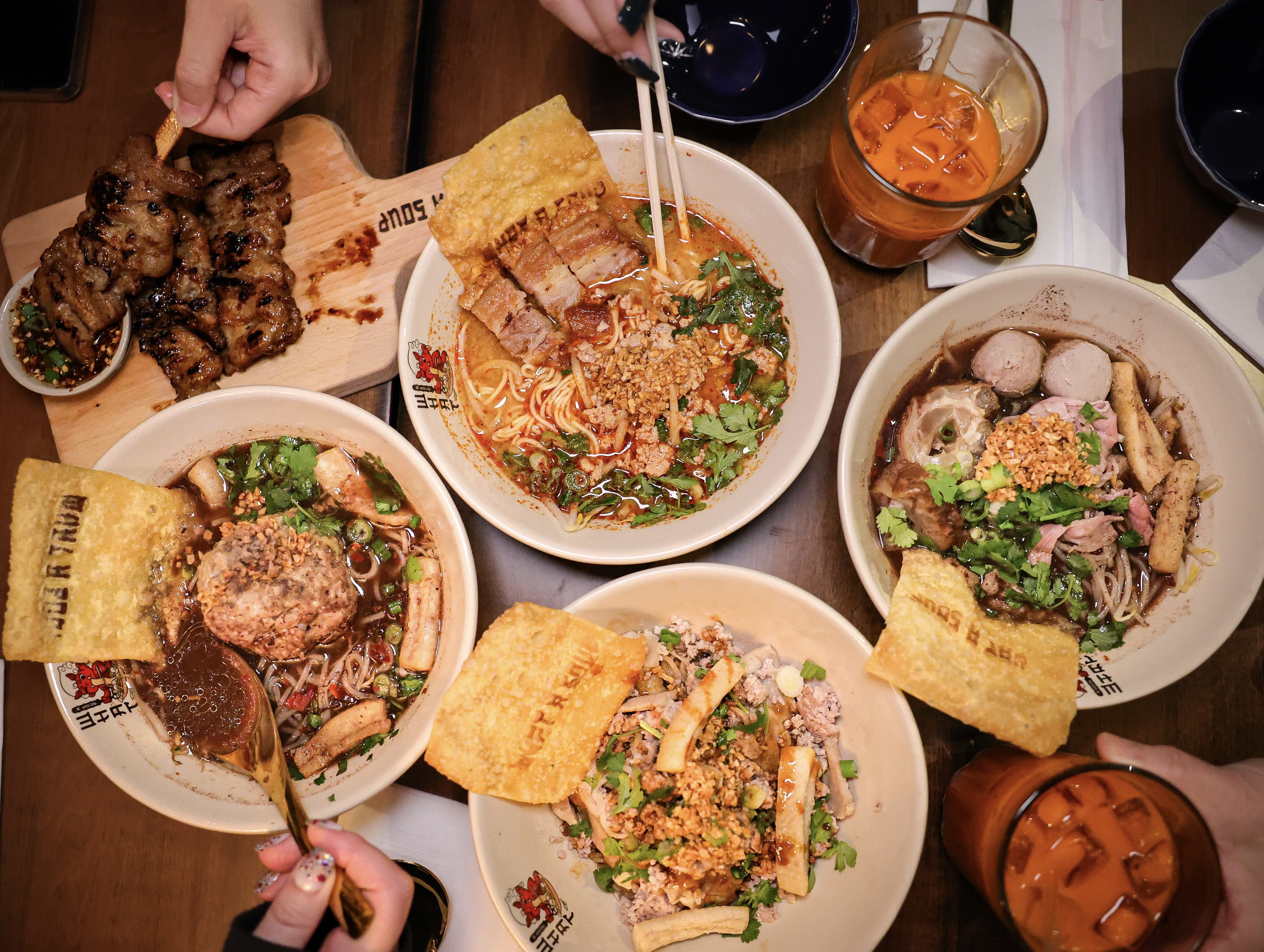 Four bowls of noodles and broth are laid out on a table with appetizers and drinks arranged around the edges.
