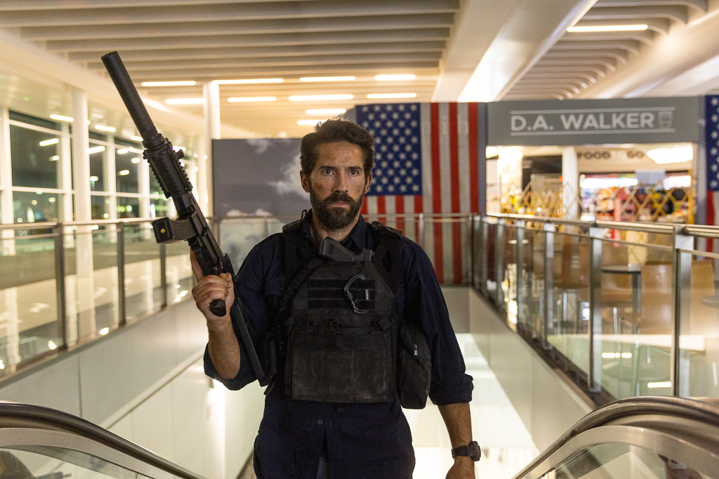 Scott Adkins wears a bulletproof vest and holds a gun while going up an airport escalator in One More Shot.