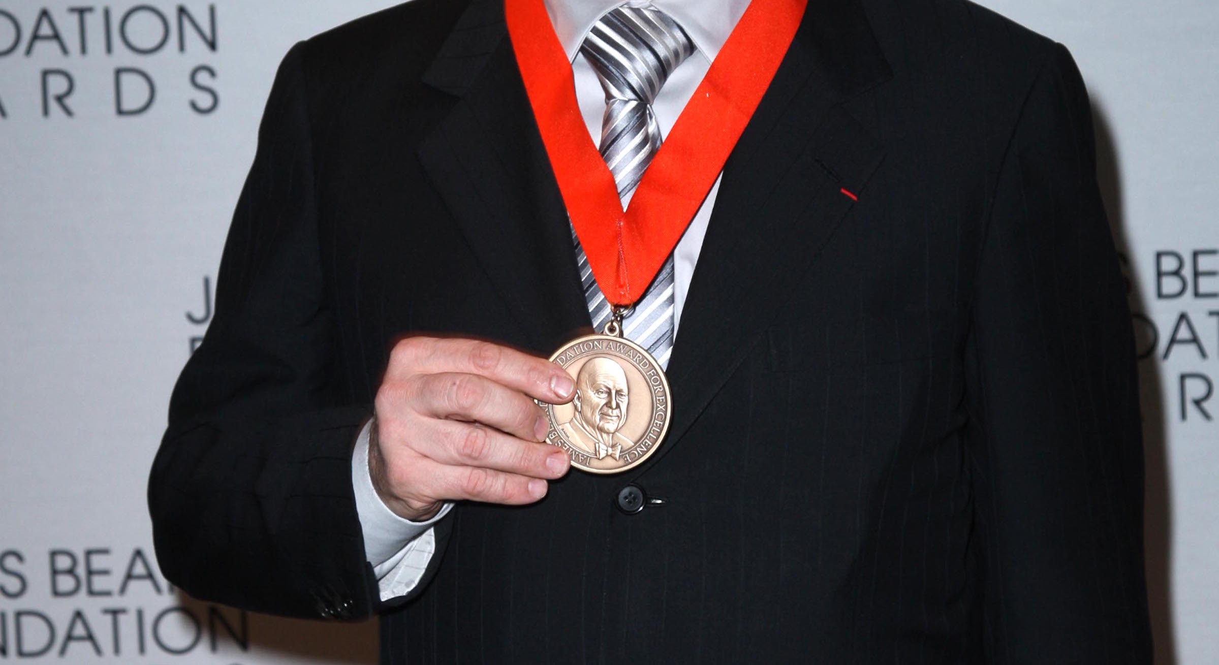 Photo shows someone holding up James Beard award medal, on ribbon around their neck.