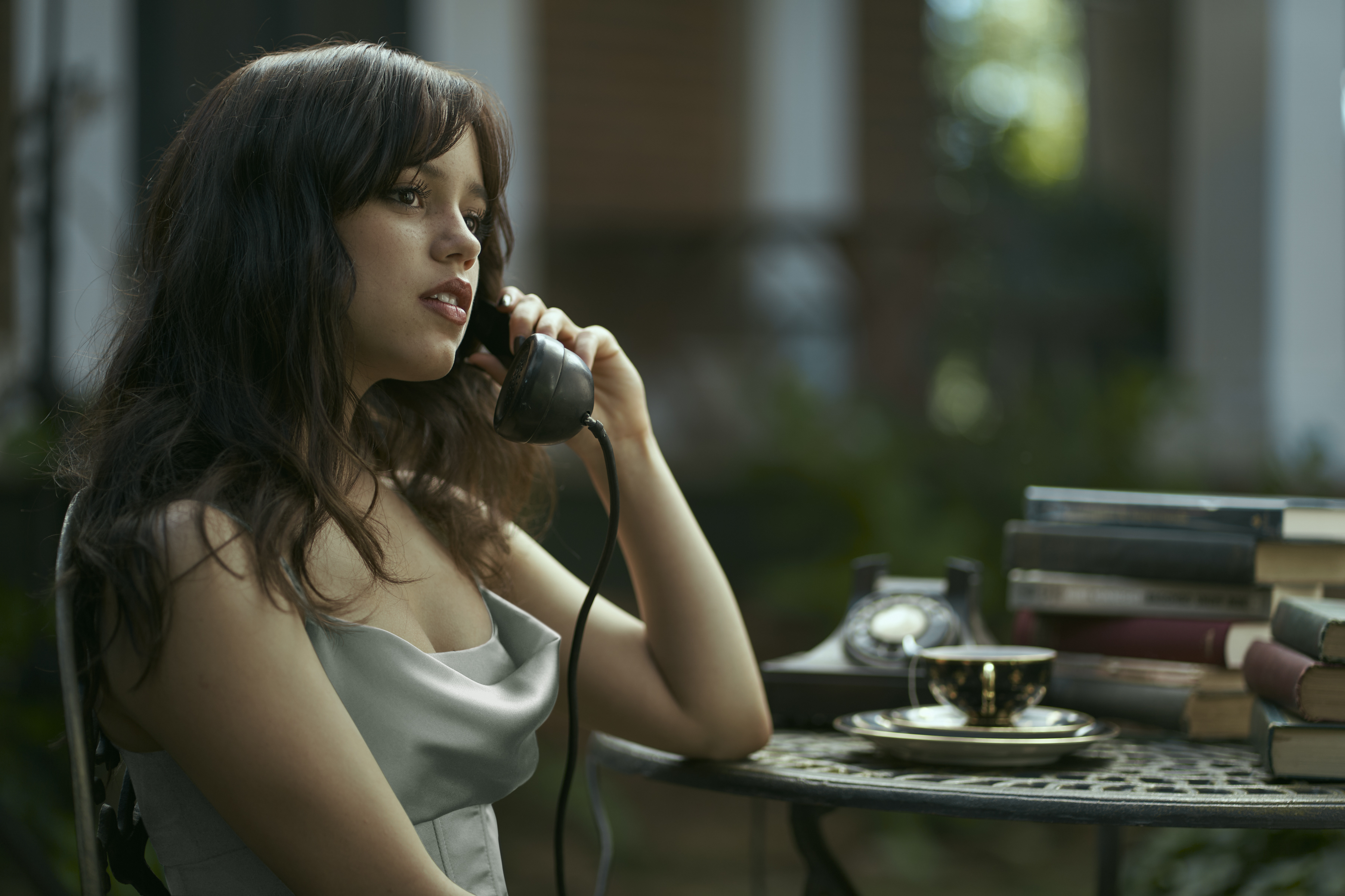 Cairo Sweet (Jenna Ortega, in a silver silk dress baring her arms and cleavage) sits at a table covered with books talking on an old-fashioned dial telephone in Miller’s Girl