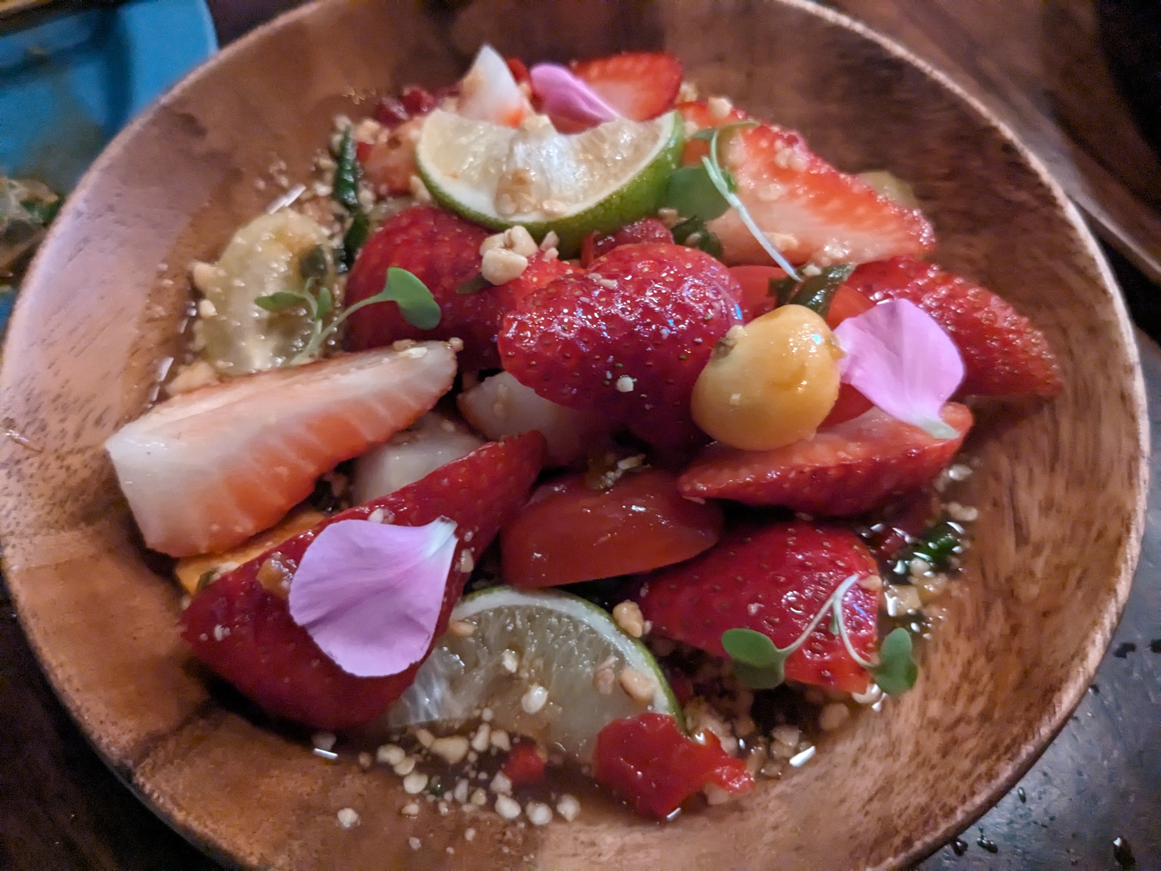 A wooden bowl with strawberries, husk cherries, and aromatics in a soupy dressing.