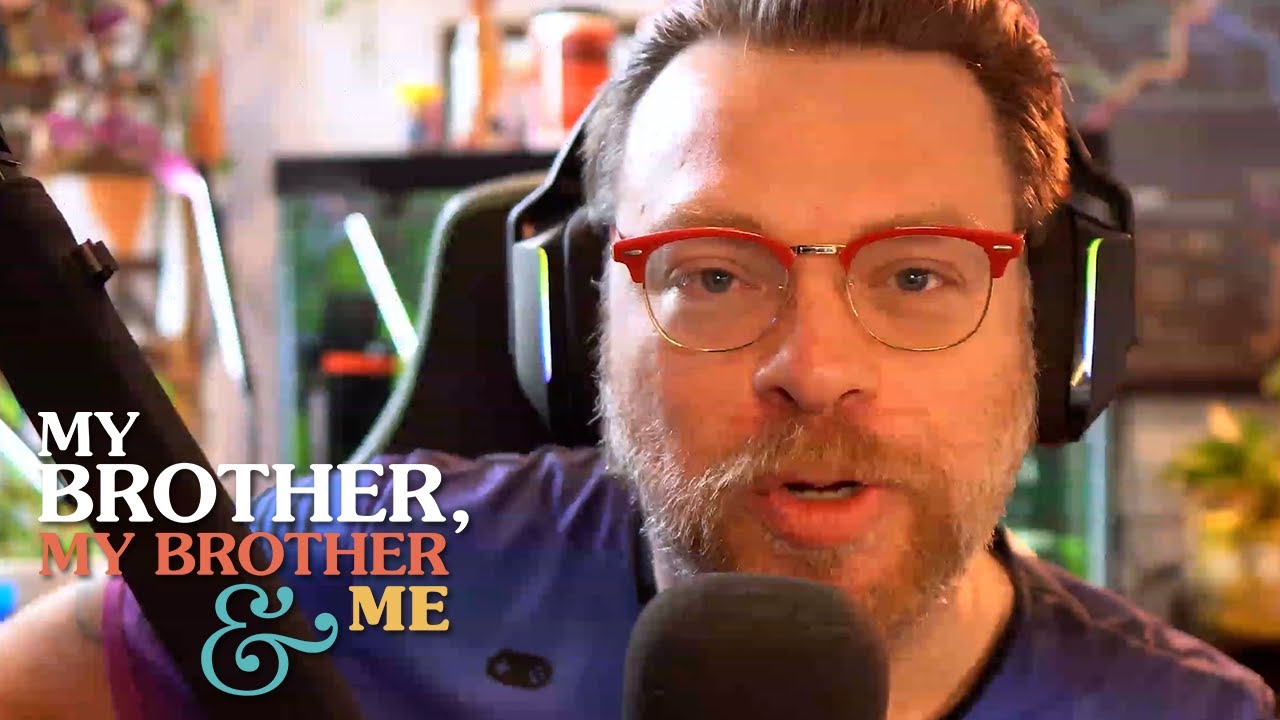 A photograph of Travis McElroy, he is seated behing a microphone and he is wearing headphones and glasses, staring directly into camera. The MBMBaM logo is superimposed in the lower left corner of the image.