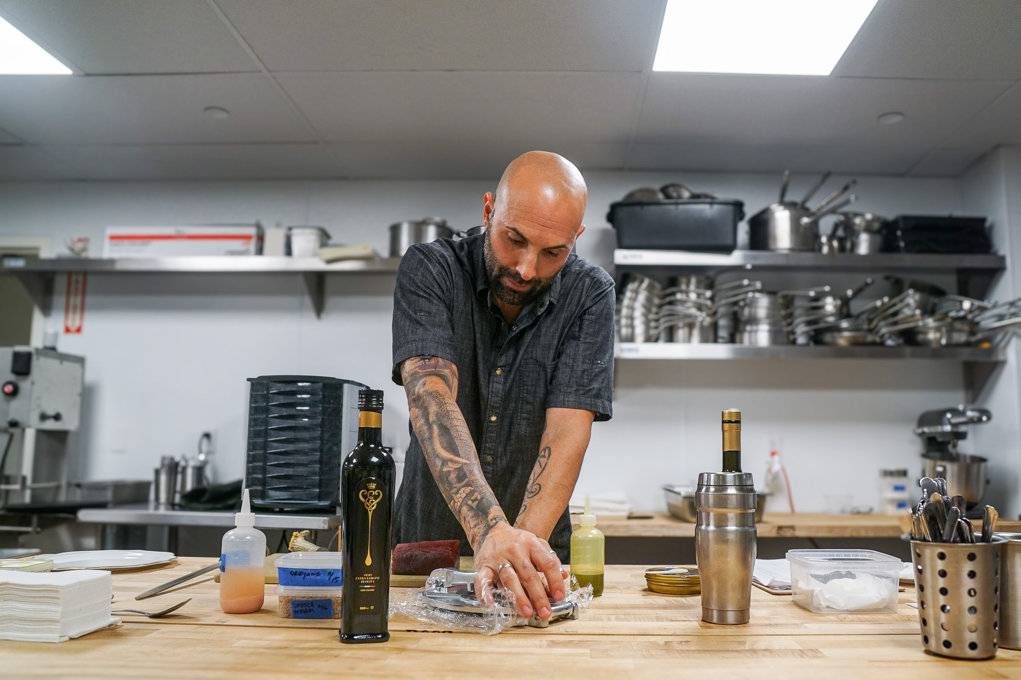 A bald male chef with tattooed arms presses a raw fish.