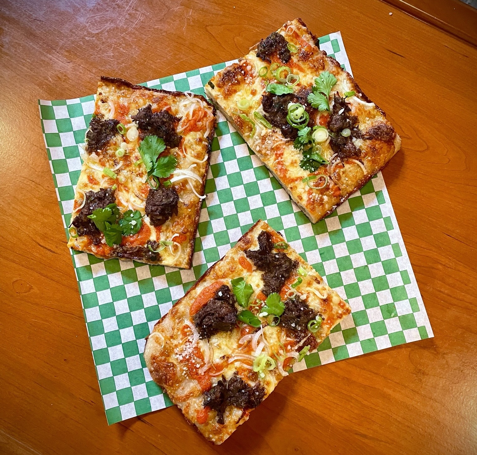 Three square slices of pizza on a green checkerboard paper with oxtail on top. All sitting on a wood table.