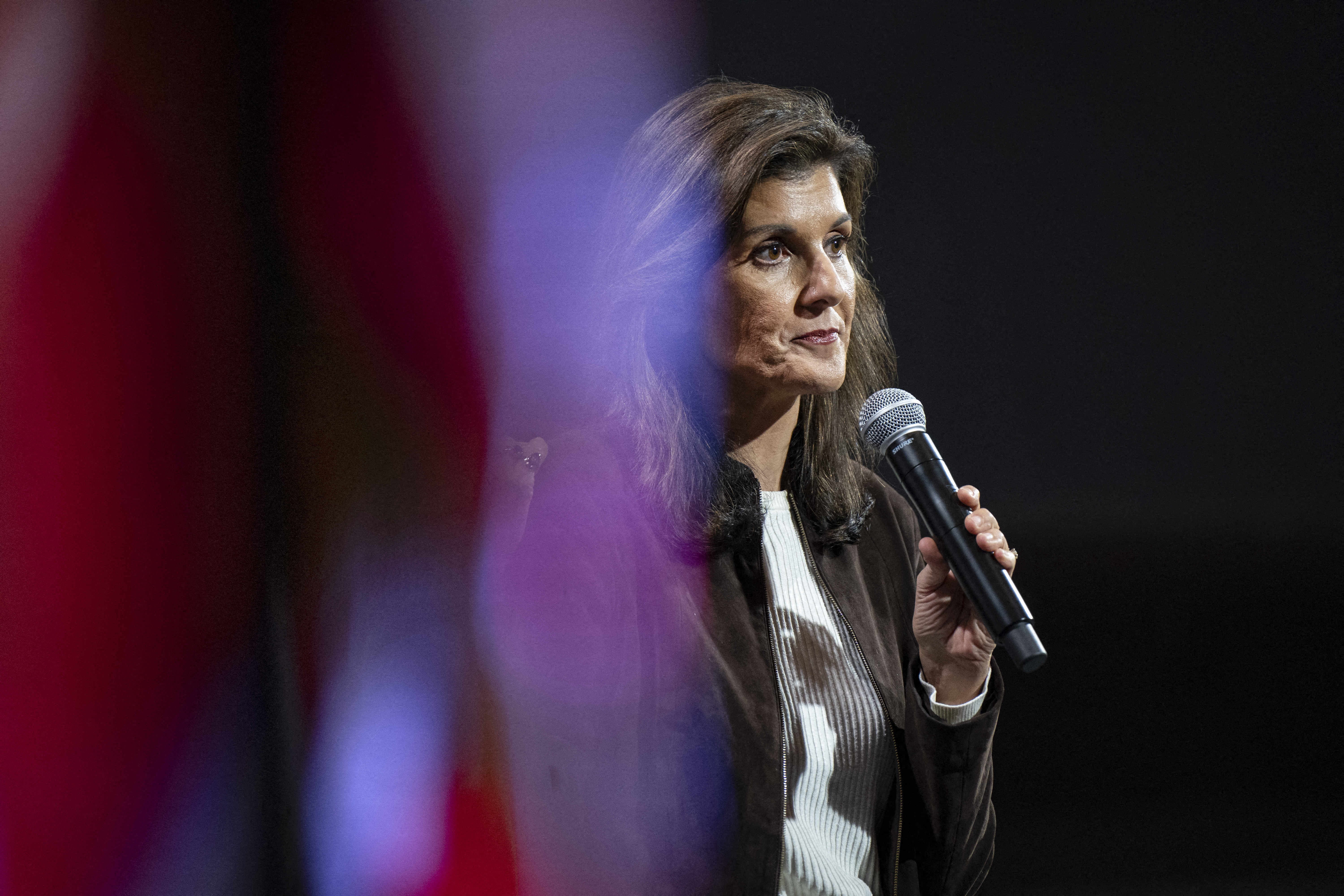Haley, in a black jacket and white shirt, looks solemn as she speaks into a microphone on a dark stage.