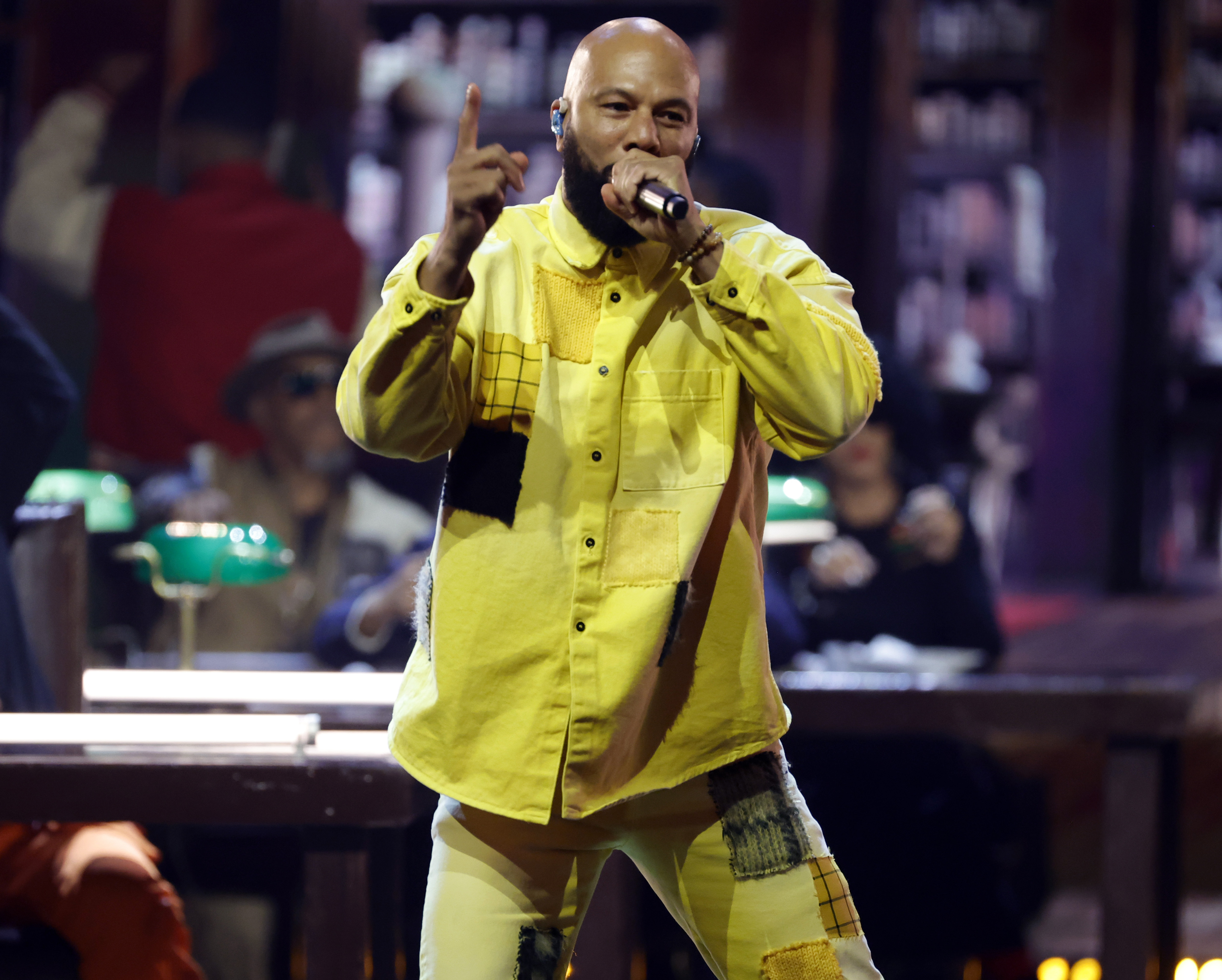 Common performs onstage on November 8, 2023, in Inglewood, California, wearing an oversized, bright yellow button-up shirt and holding a microphone. 