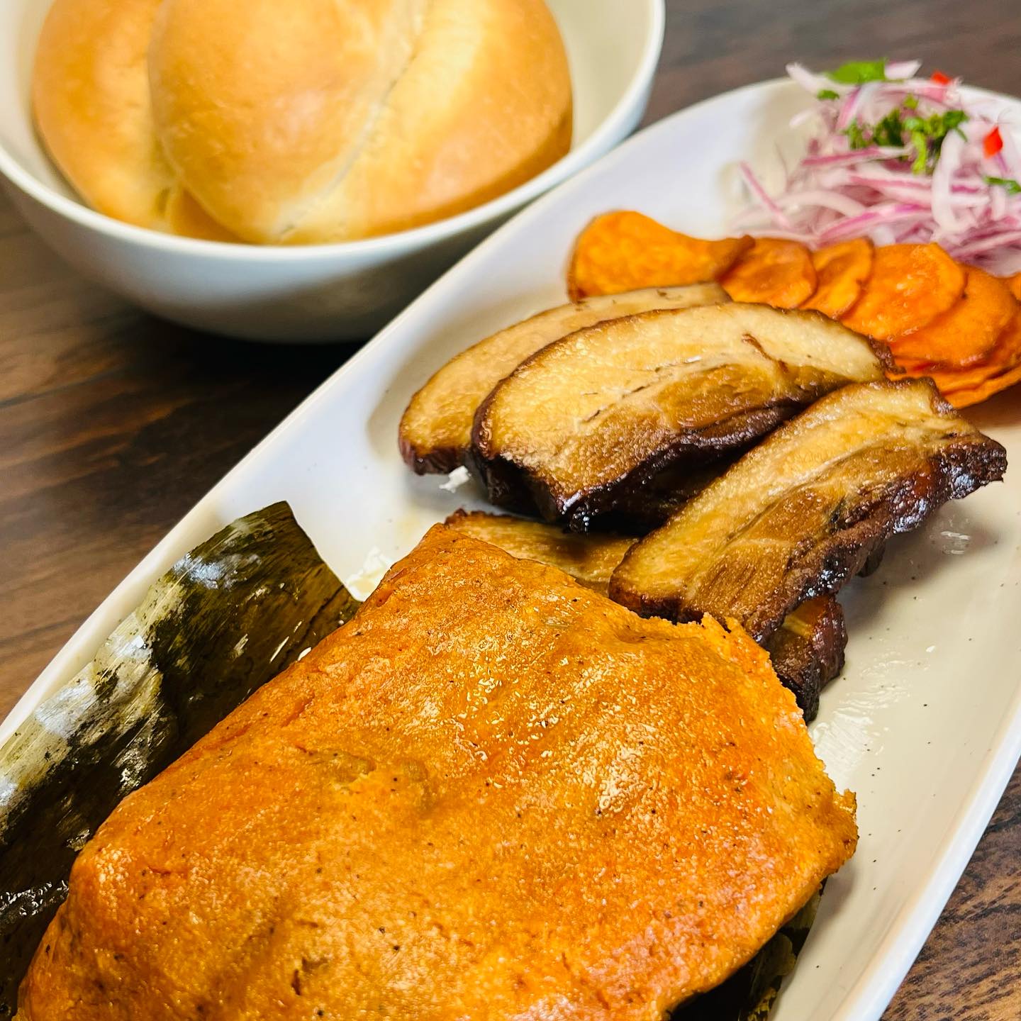 A white platter with French bread, Peruvian chicharron, sweet potato with criolla sauce, and a tamal