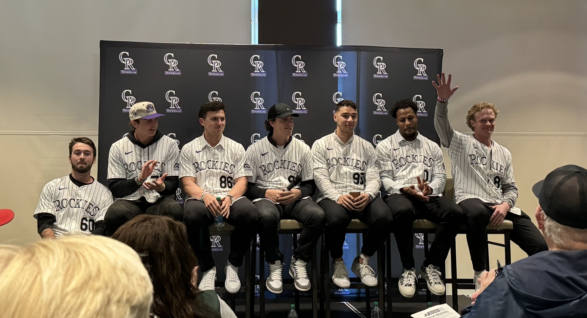 Sterlin Thompson, Zac Veen, Drew Romo, Chase Dollander, Gabriel Hughes, Jaden Hill, and Case Williams on the stage at RockiesFest. Each is wearing a purple-pinstripes Rockies jersey and sitting on a tall stool. They’re also wearing some very nice trainers.