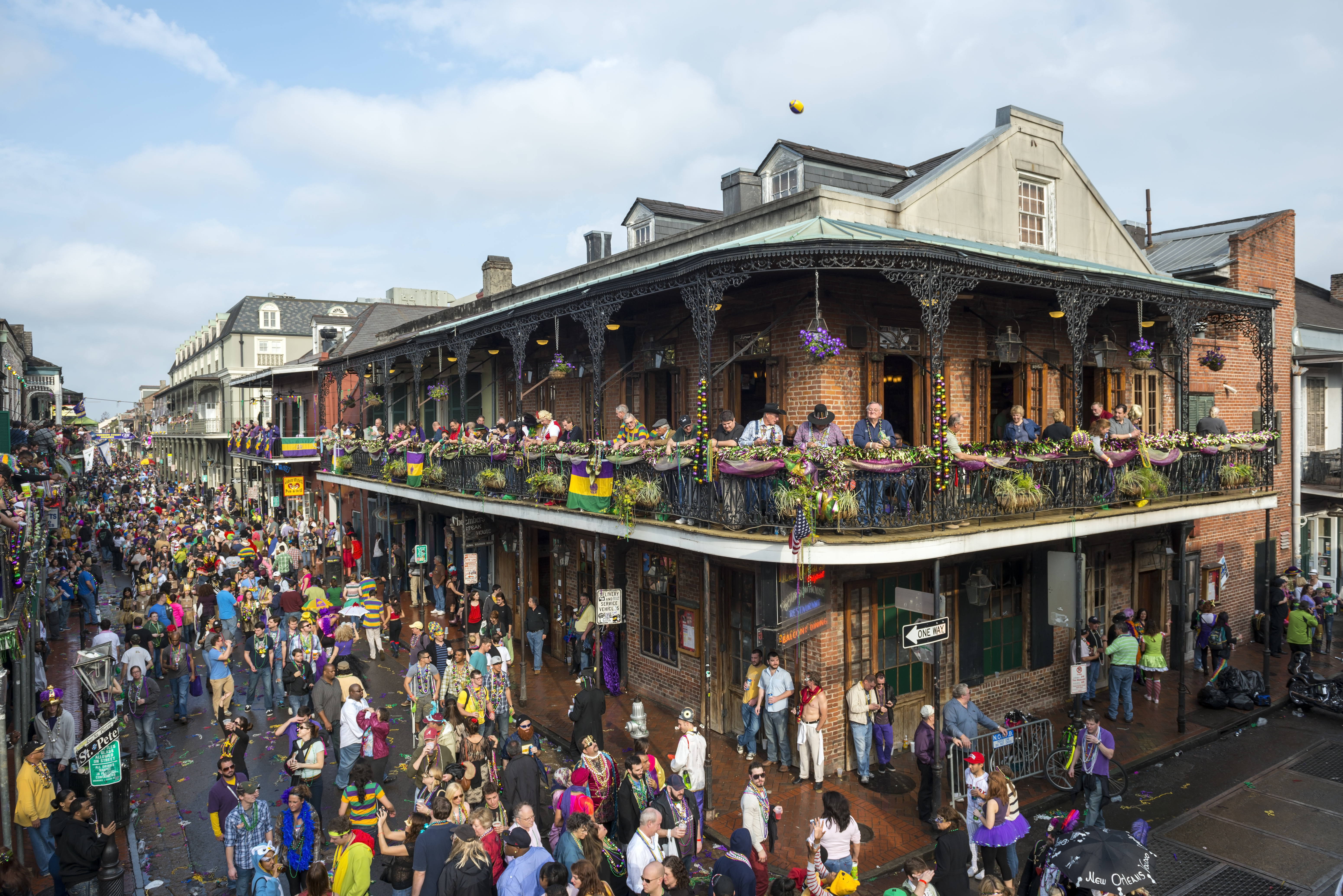 A two-story building with wraparound porches on both levels; crows of peopled around it on the street, celebrating Mardi Gras. 