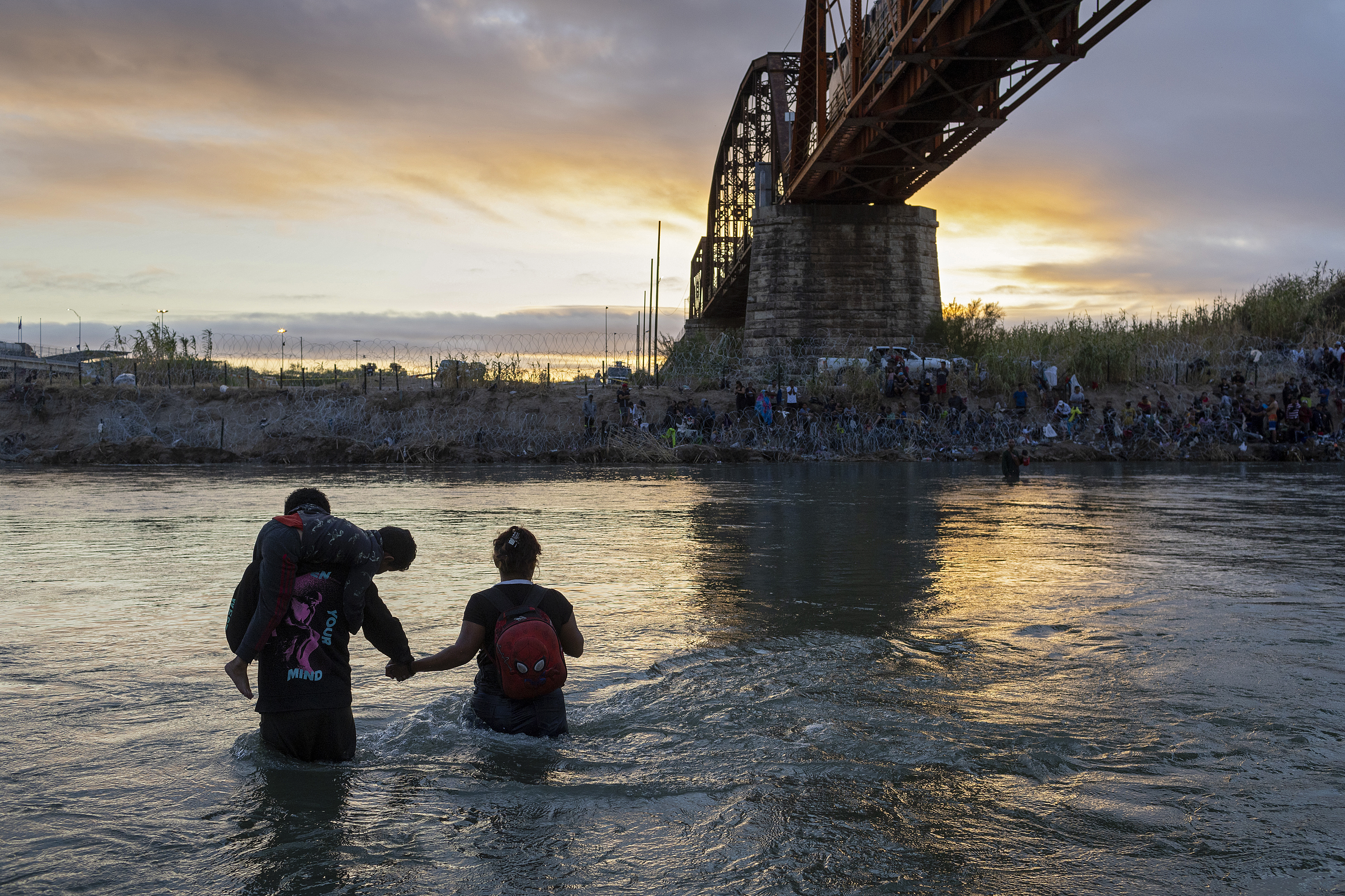 People holding hands while wading across a wide river below a bridge, at dusk.