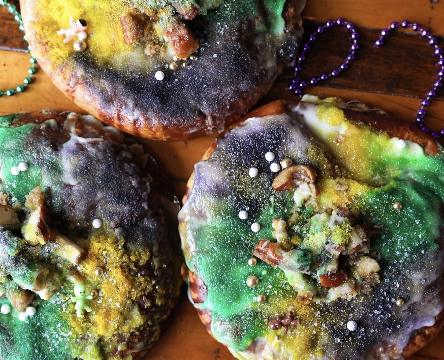 Three king cakes topped with green, white, yellow, and purple frosting sitting on a wooden surface with purple and green beaded necklaces surrounding it