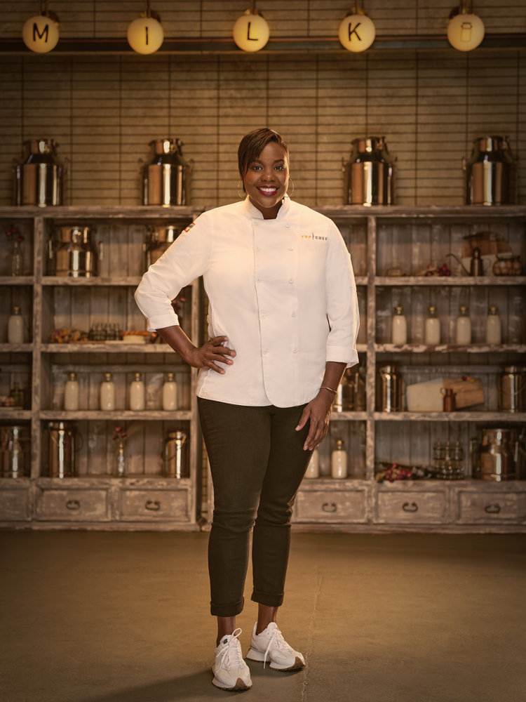 A woman in chef whites stands posed in front of a pantry full of staple foods.