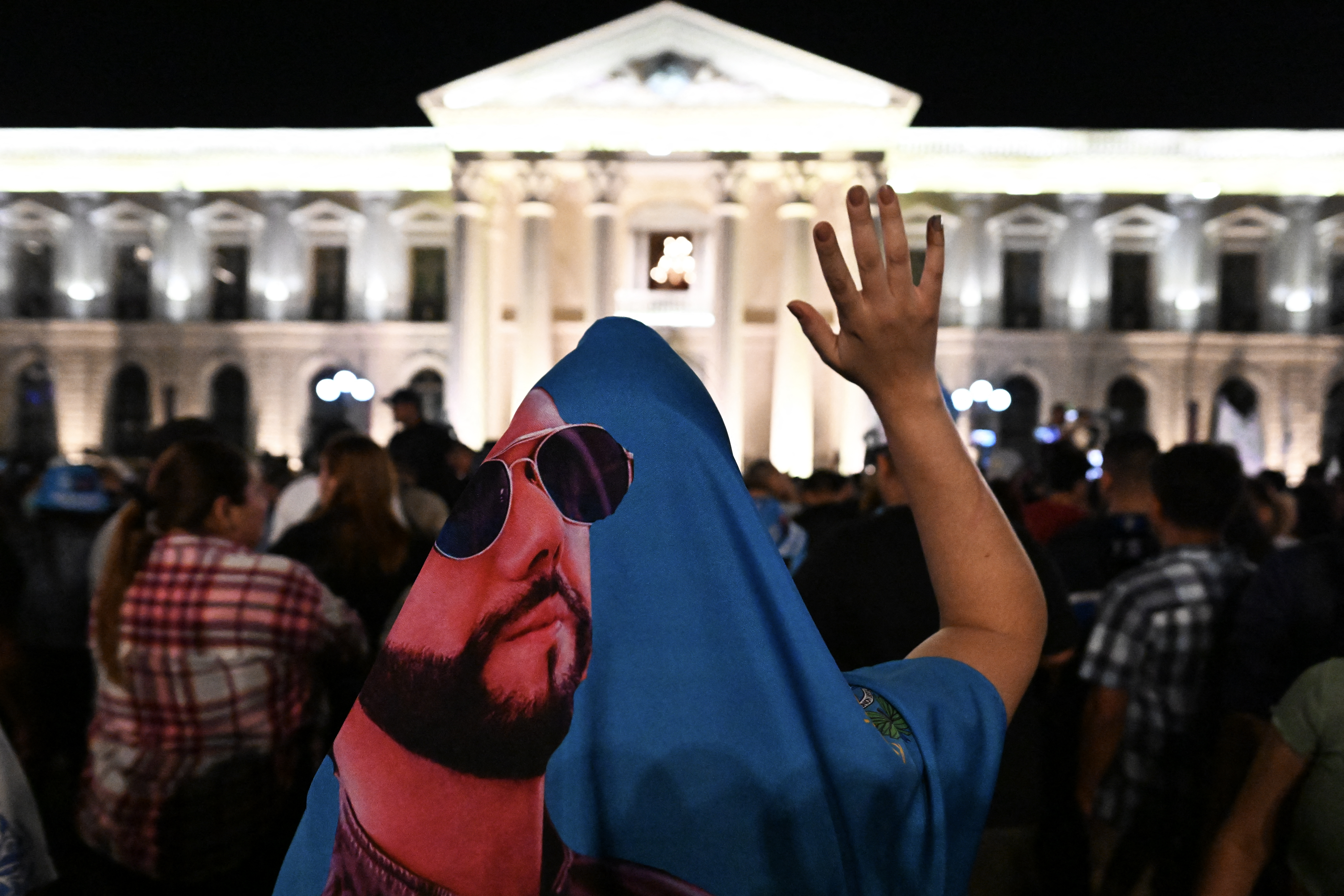 Bukele supporters gather outside the National Palace in San Salvador at night. The person closest to the camera is draped in a printed image of Bukele wearing sunglasses.