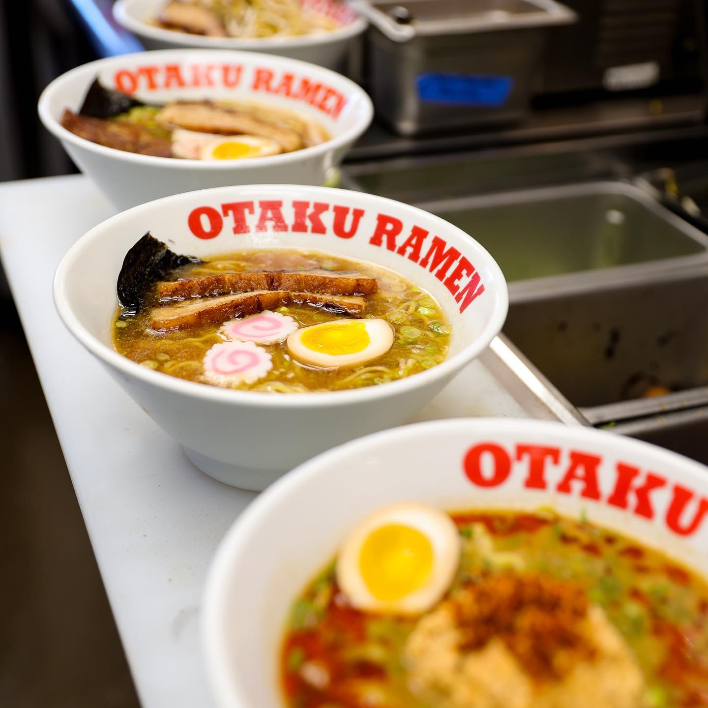 Three bowls of ramen lined up in a row. The white bowls are emblazoned with the Otaku Ramen logo in red