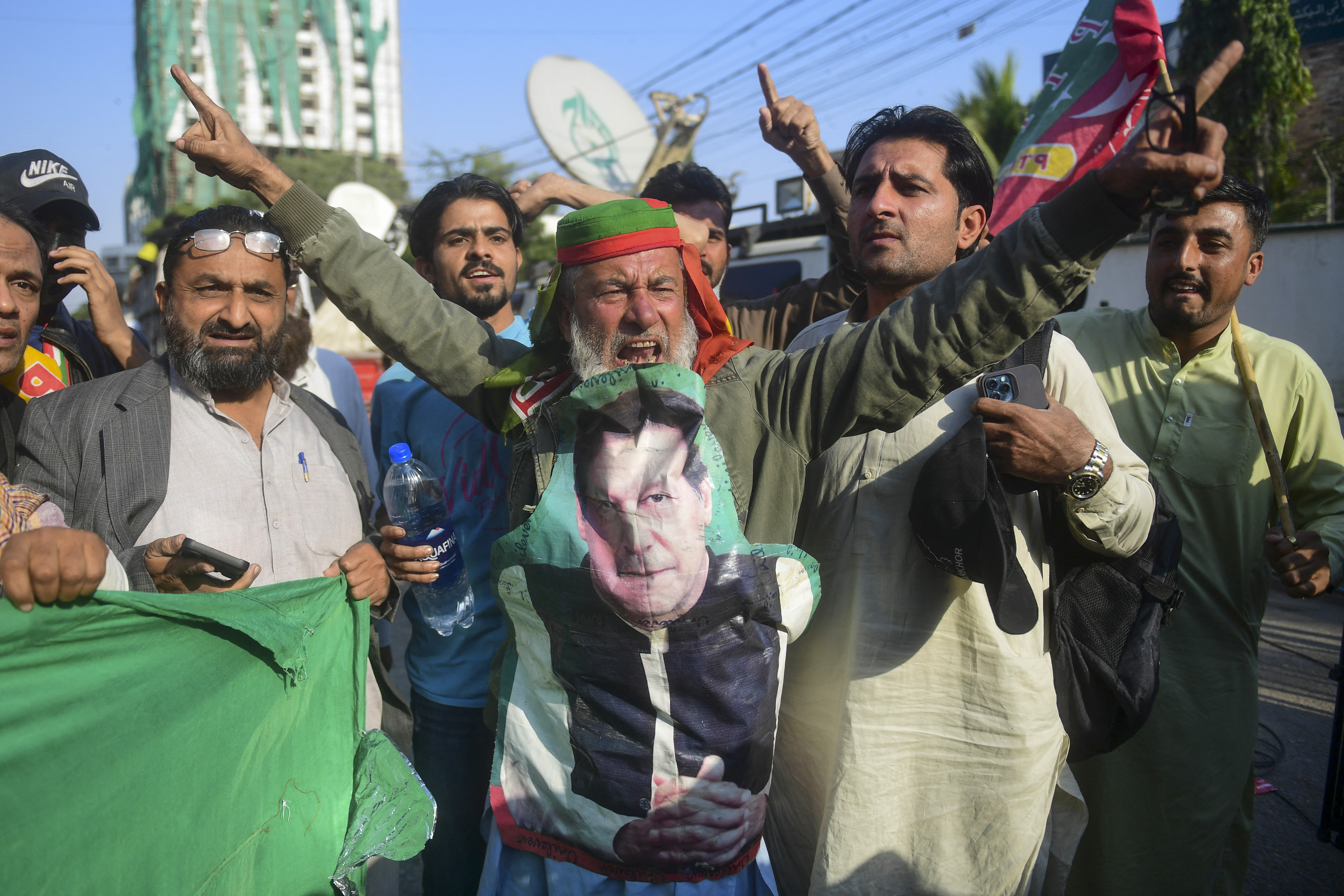 An elderly Pakistani man with a long white beard wears a giant cutout of Khan on his chest and raises his arms in victory, surrounding by jubilant, cheering men pointing at the blue sky.
