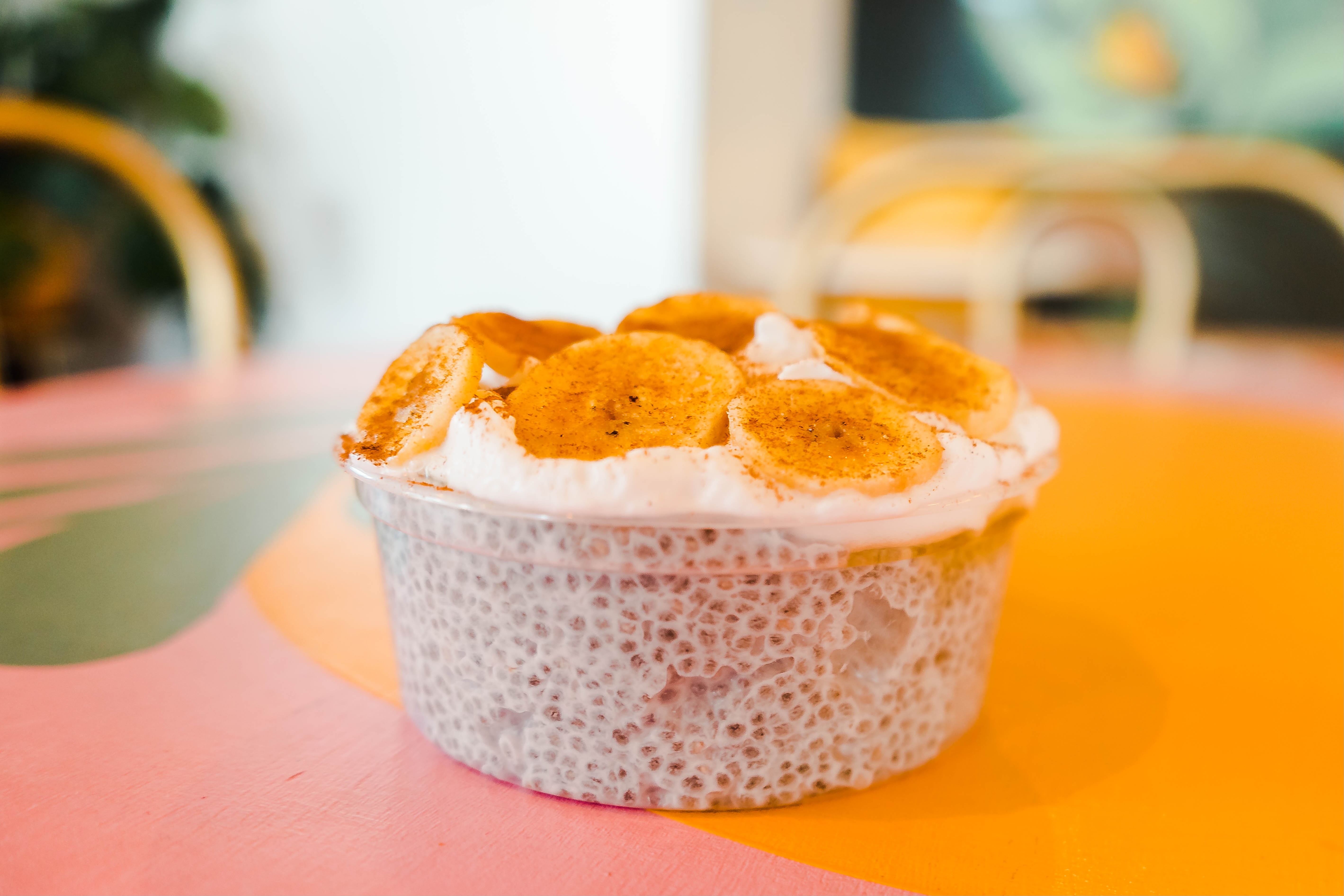 A plastic dish of chia seed pudding topped with coconut cream and bananas sitting on a pink and orange tabletop. 