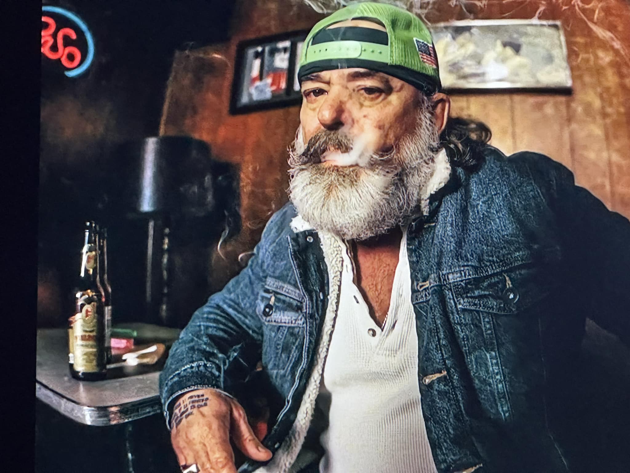 A man with a beard in a backward baseball cap sits in a bar smoking.