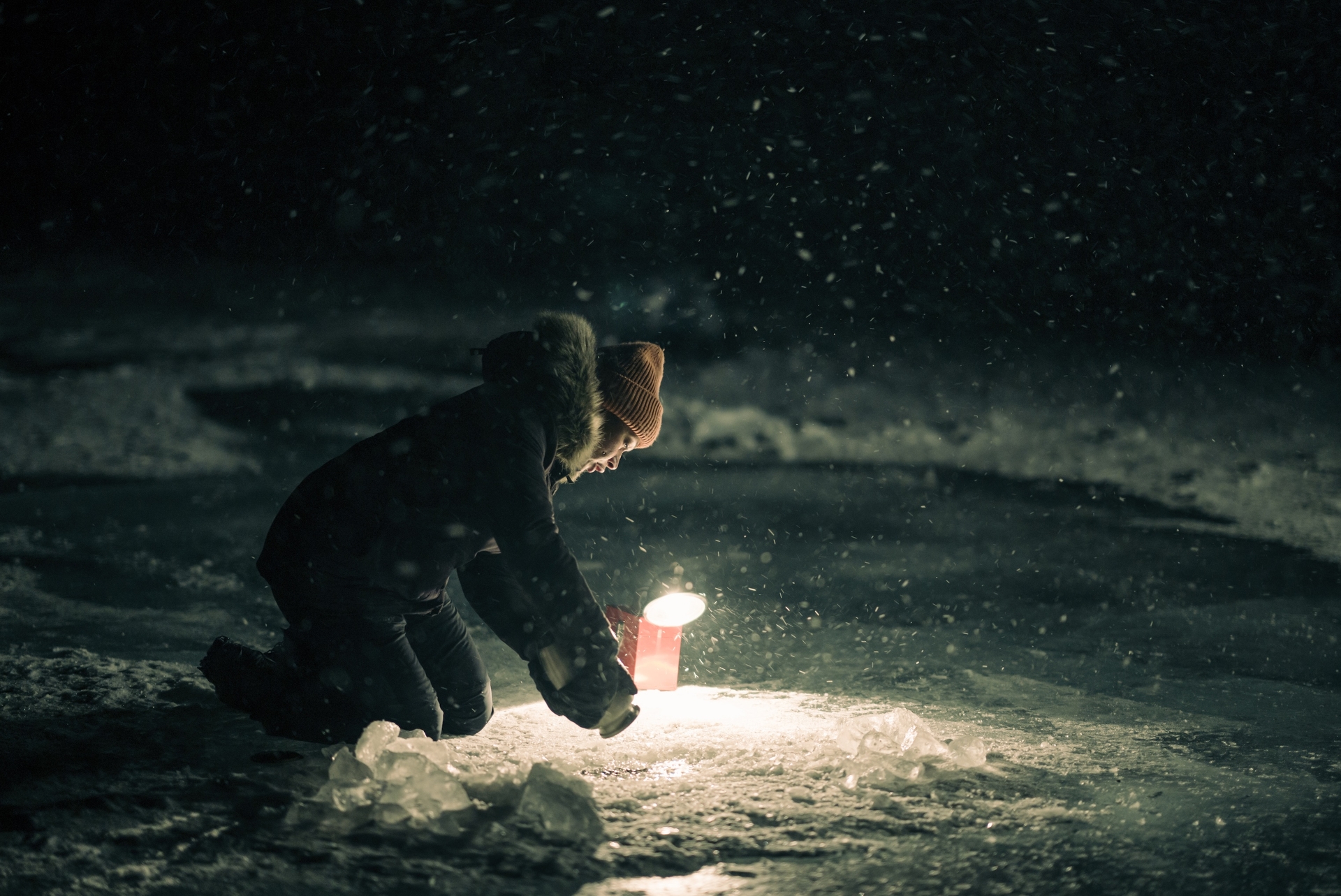 A woman holds a flashlight while kneeling, the light showing ice-covered water.