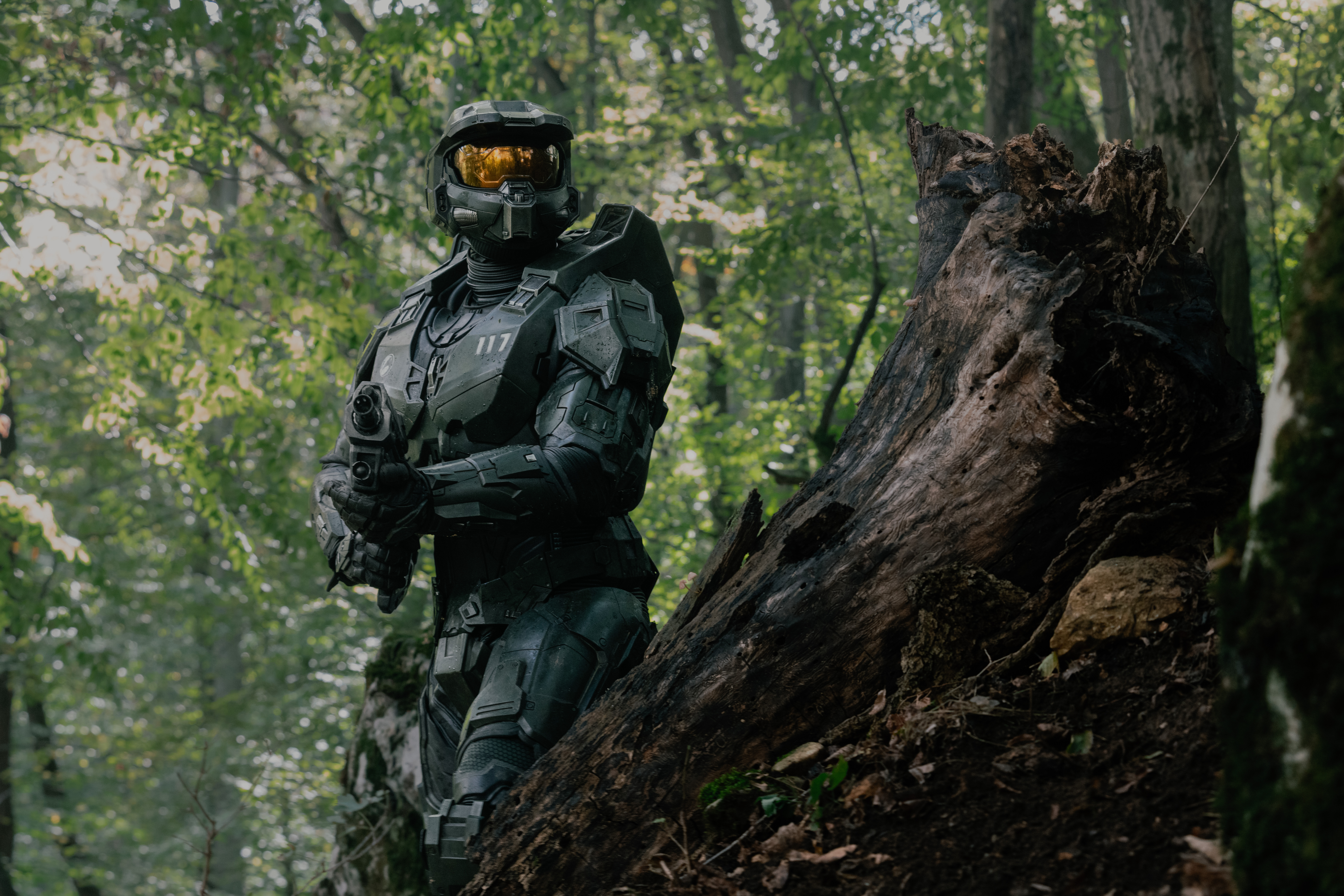 Pablo Schreiber in the Master Chief armor holding an assault rifle and walking through the woods with a tree stump in front of him in Halo season 2 