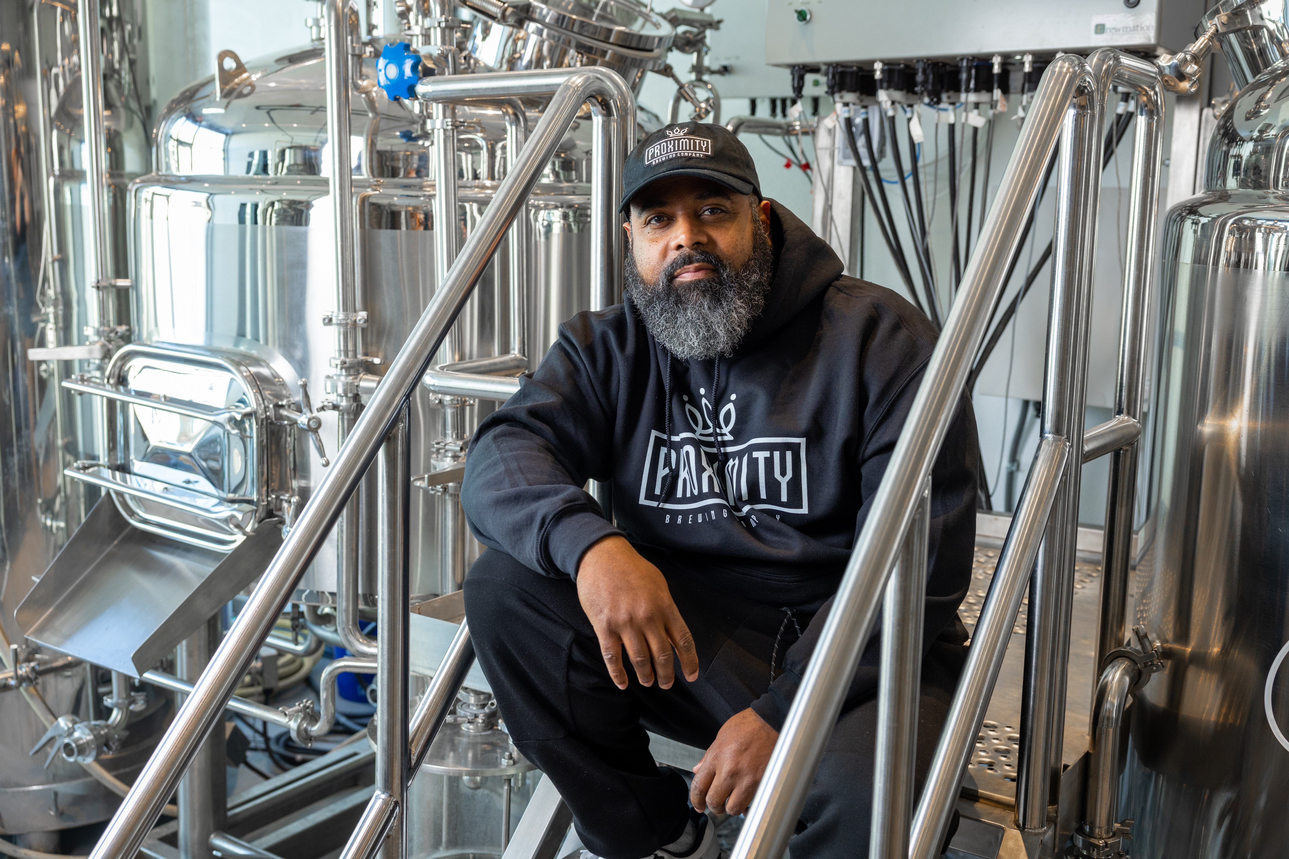 A Black man with a beard, wearing a hat, sitting in front of stainless steel brewing equipment.