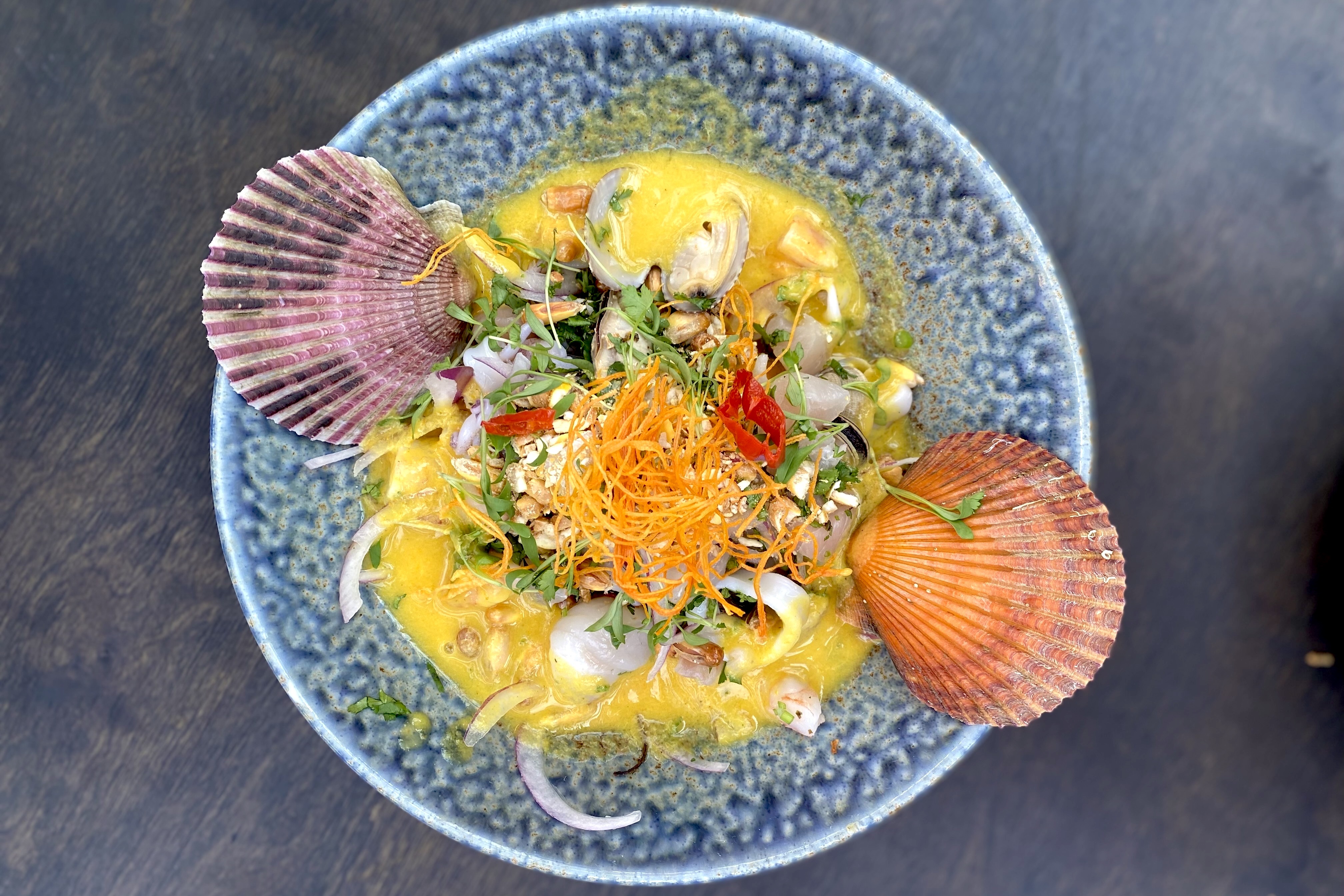 A blue bowl filled with ceviche in a yellow sauce with two seashell accents, one orange and one purple. 