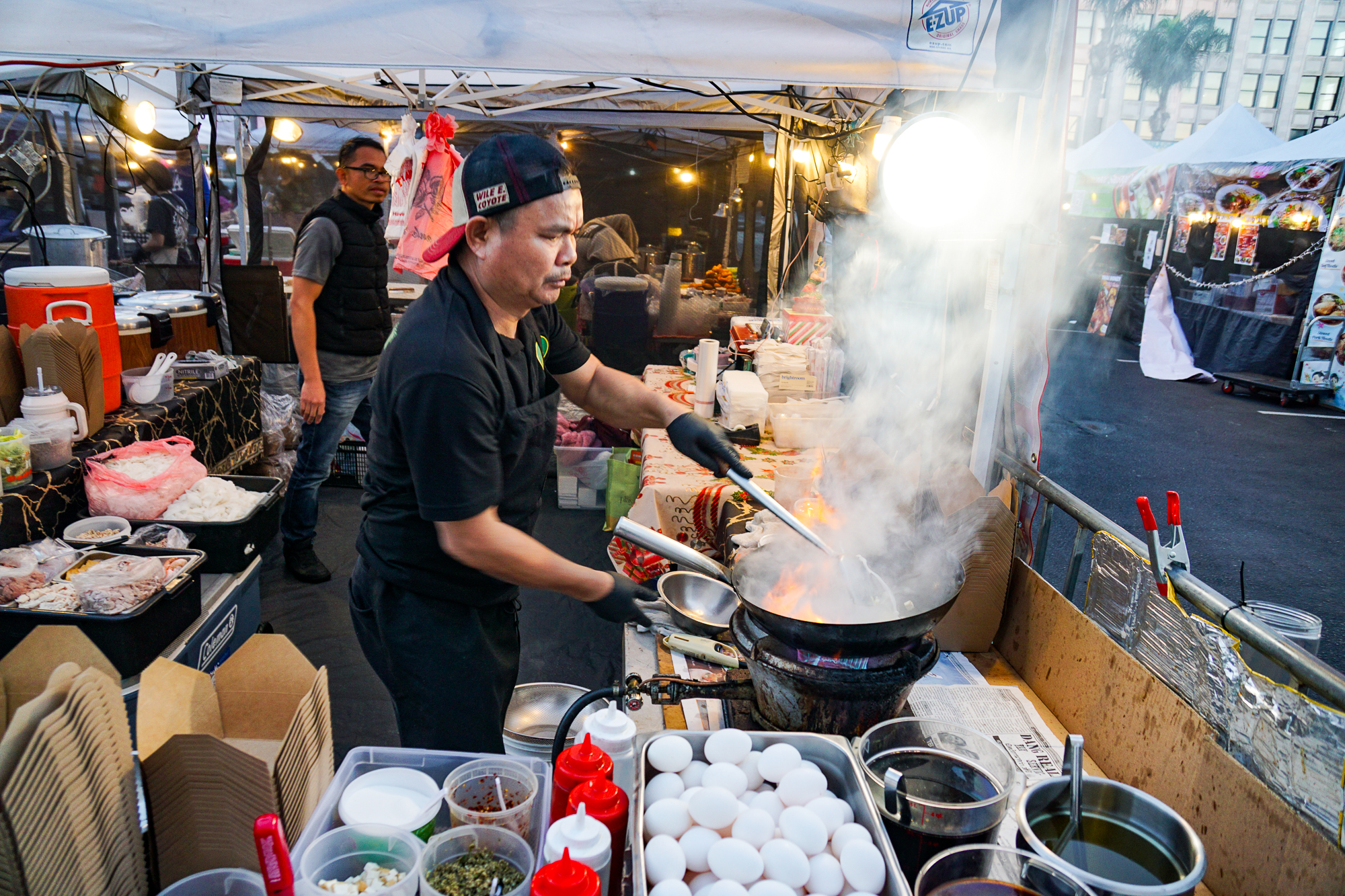 A man wearing a black hat, shirt, and pants in front of a steaming hot wok with a lot of smoke at At Siam.
