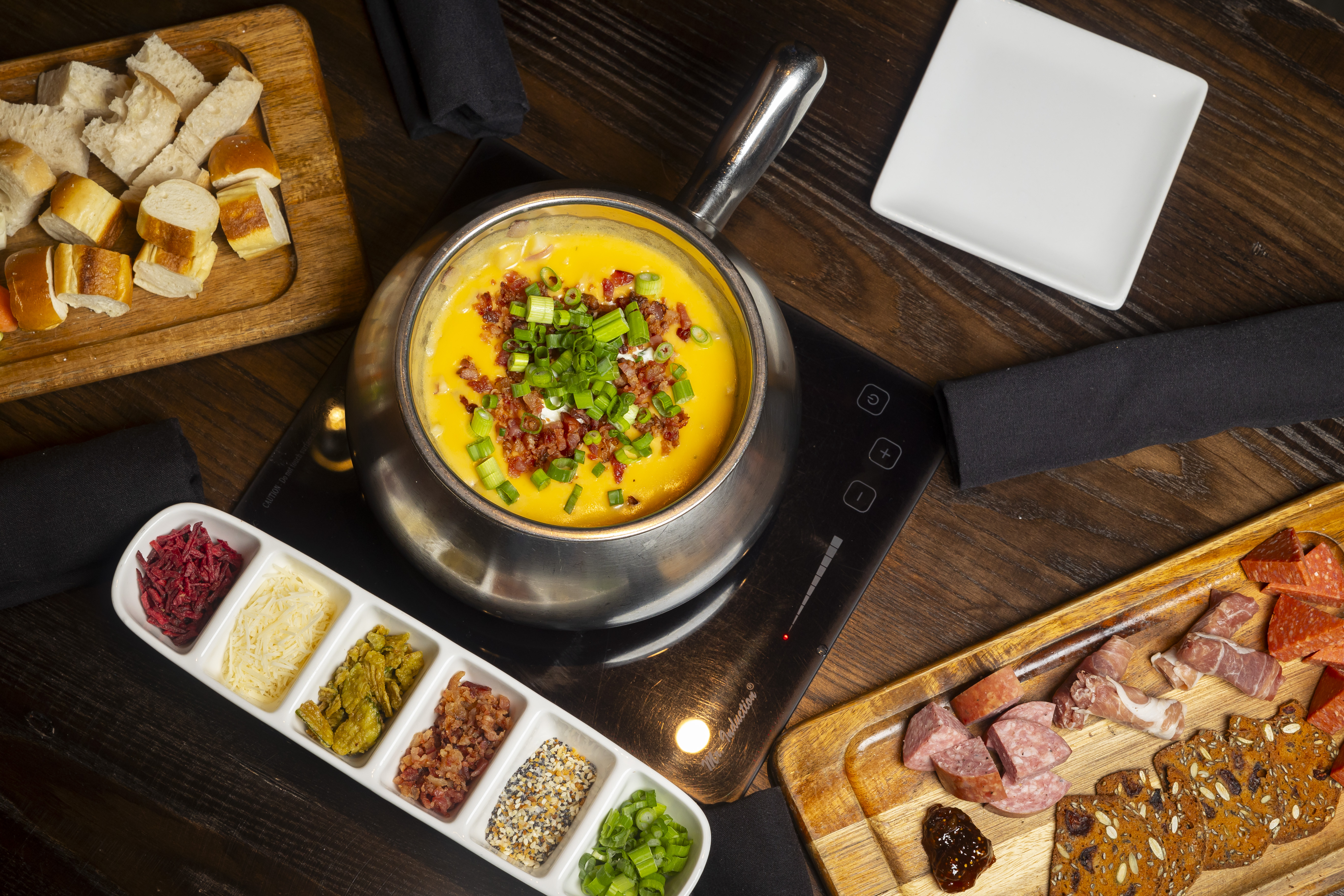 A fondue pot filled with cheese atop a table, alongside a dish of fondue toppings and cutting boards ladenwith bread and chunks of meat.