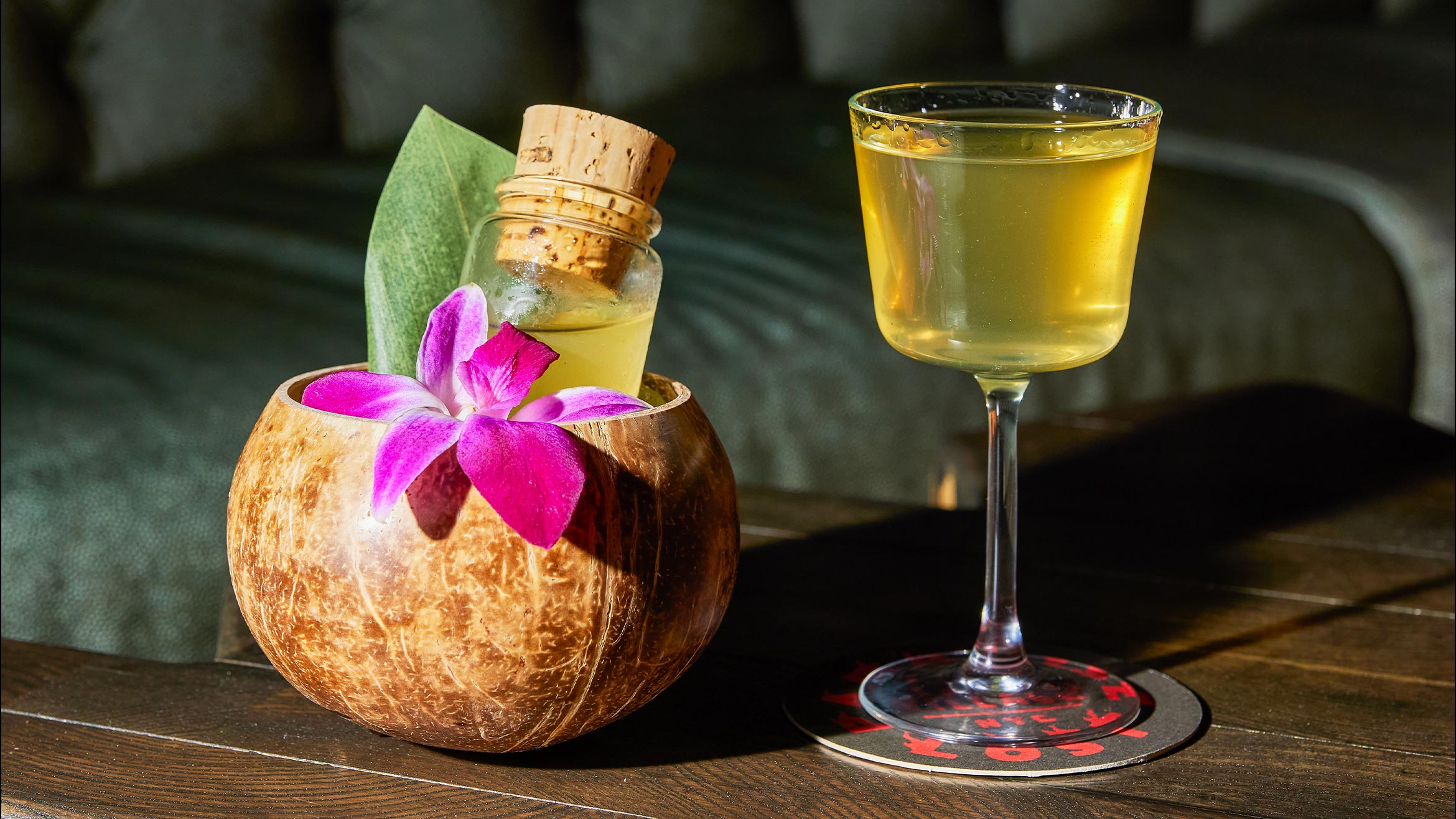 A half coconut shell holding a bottle of liquid closed with a cork and a floral garnish alongside a small, but full, cocktail glass