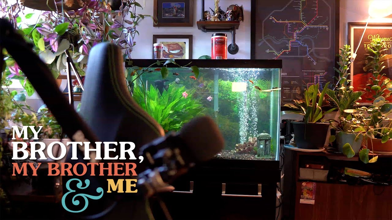A photo of Travis’ office, with him notably absent. His microphone, chair, and the fish tank behind the chair are all visible. The MBMBaM logo is superimposed in the lower left corner.