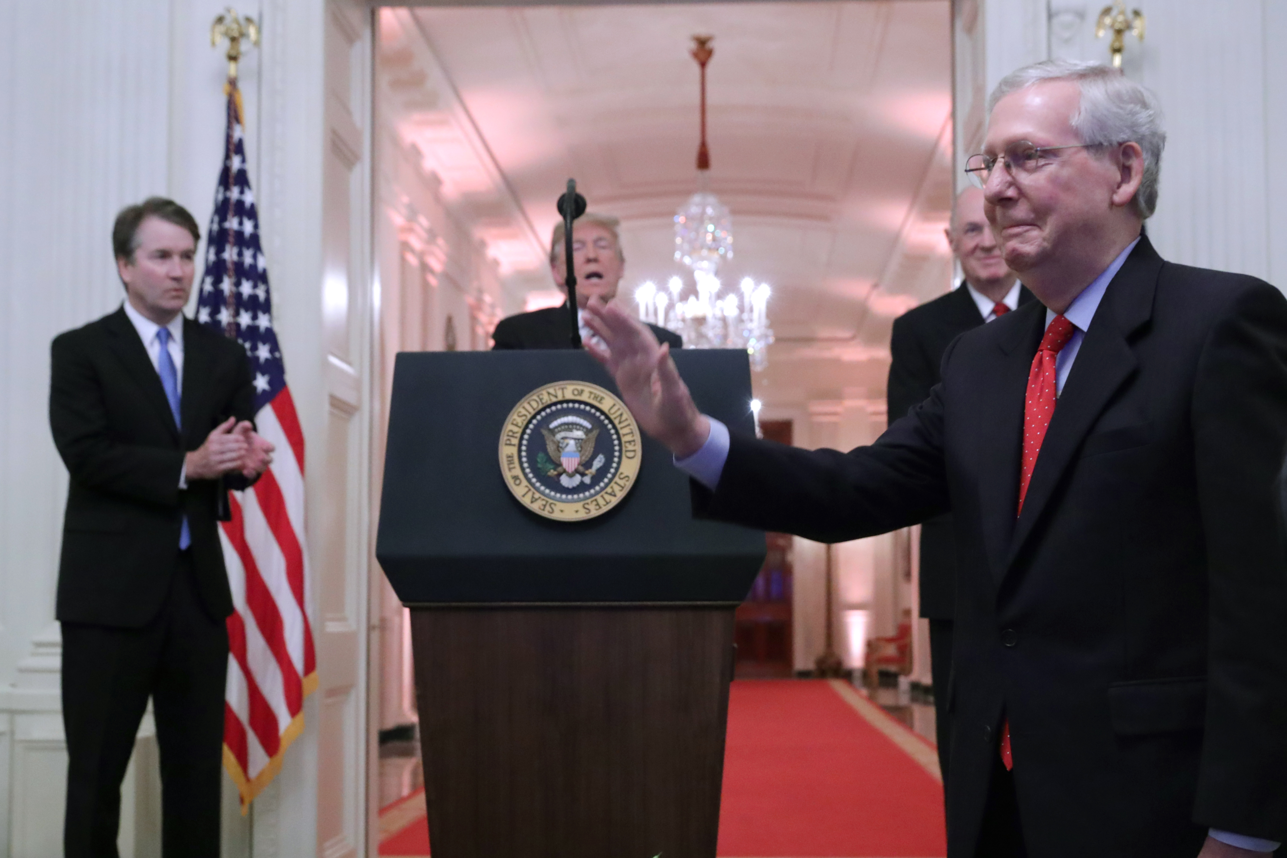 McConnell waves in front of a podium, with Trump and Kavanaugh in the background at the White House.