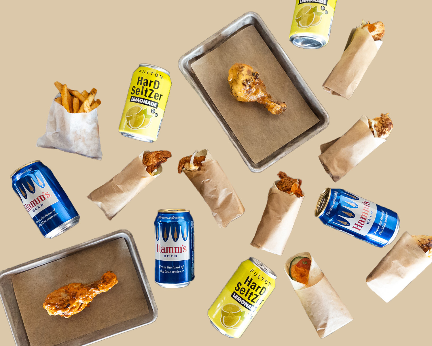 An assortment of chicken snack wraps in brown paper, drumsticks on small trays lined with brown paper, French fries in white paper bag, and cans of Hamm’s beer and hard lemonade cut out and arranged on a tan background. 