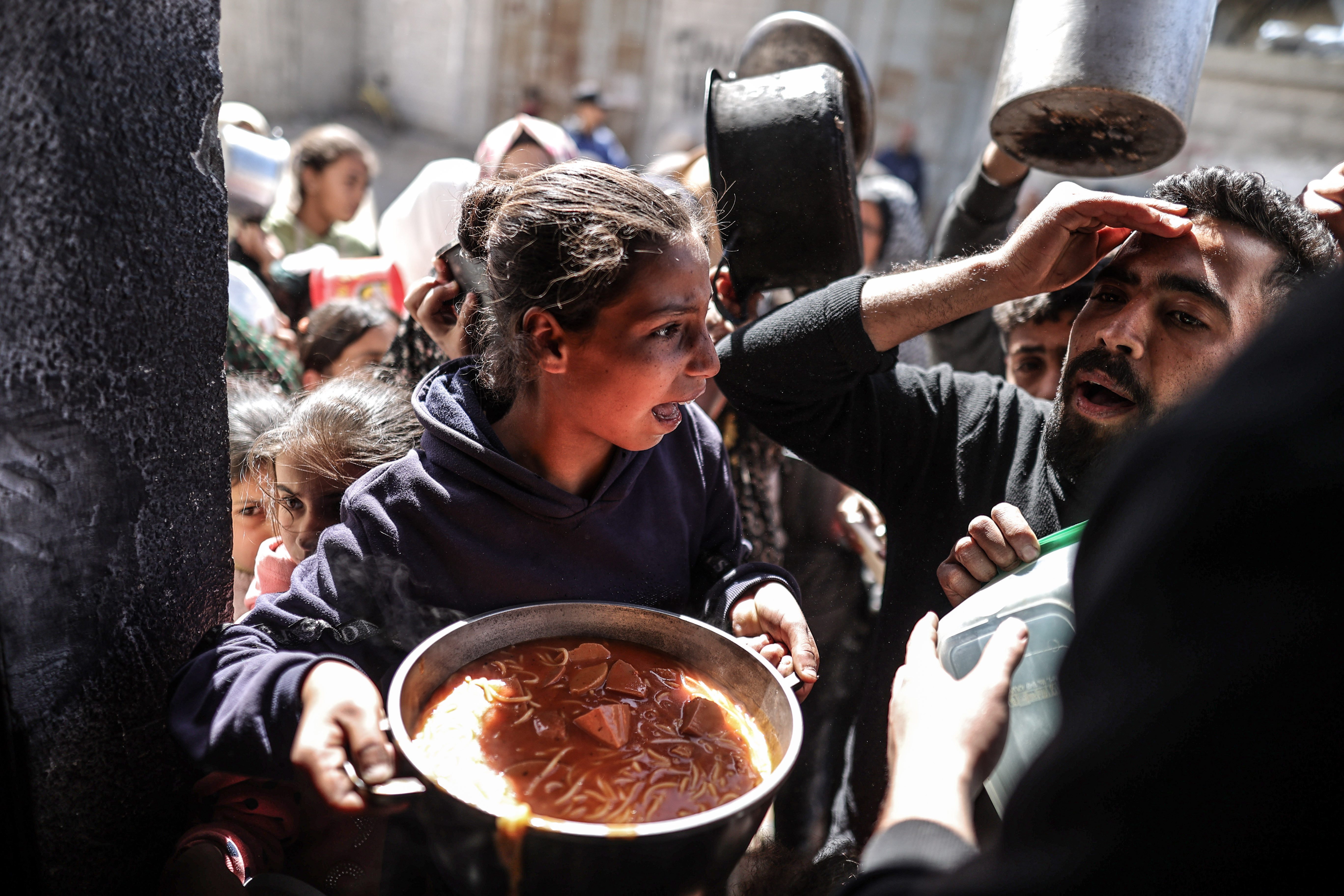 A girl holds a pot of food while being jostled by a crowd.