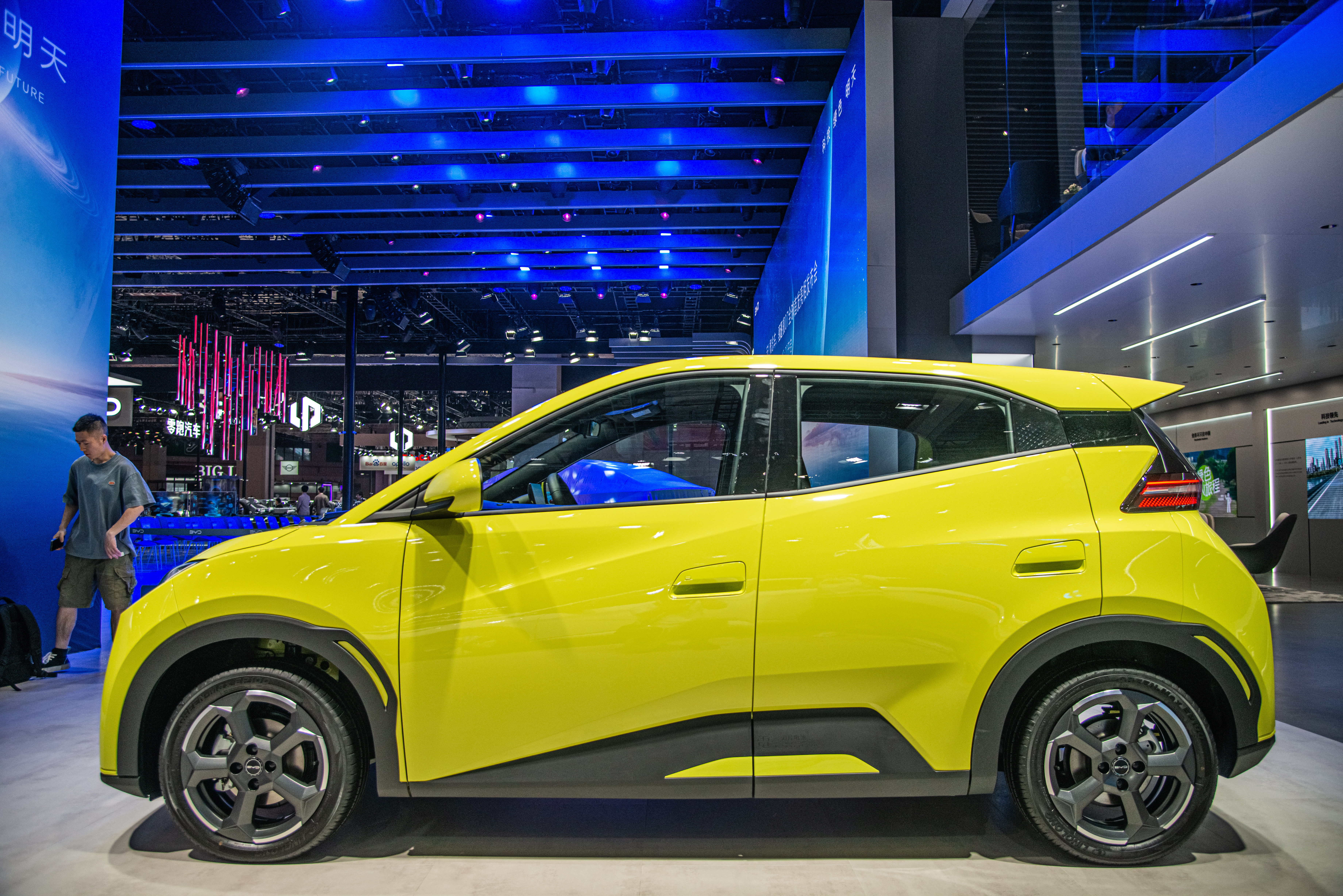 A squat yellow hybrid car on display in a large room with a high, blue-lit ceiling. 
